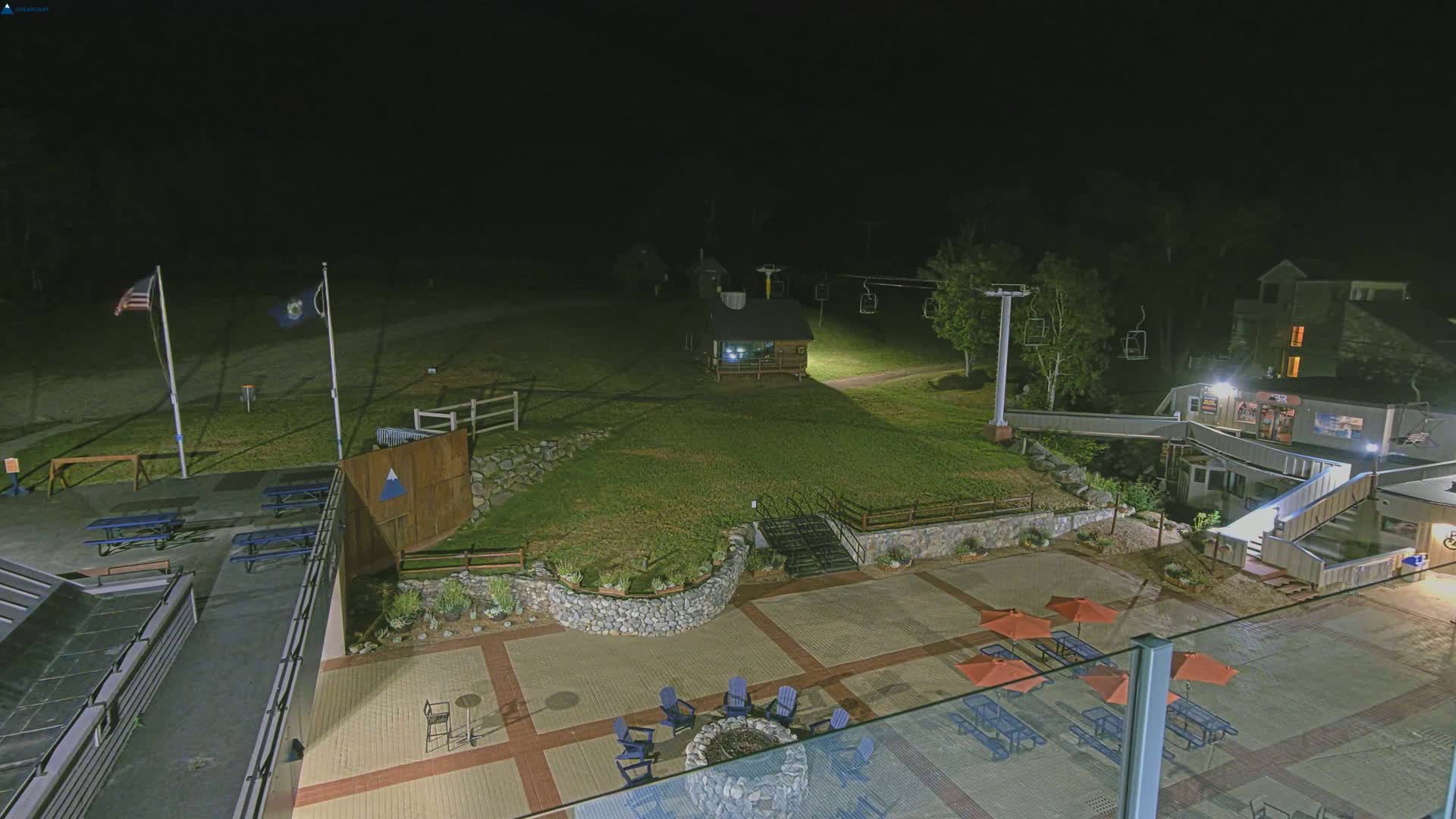A nighttime view of a ski resort base area, featuring a paved patio with outdoor seating and fire pit, a small building, and a ski lift in the background under dark, clear skies.