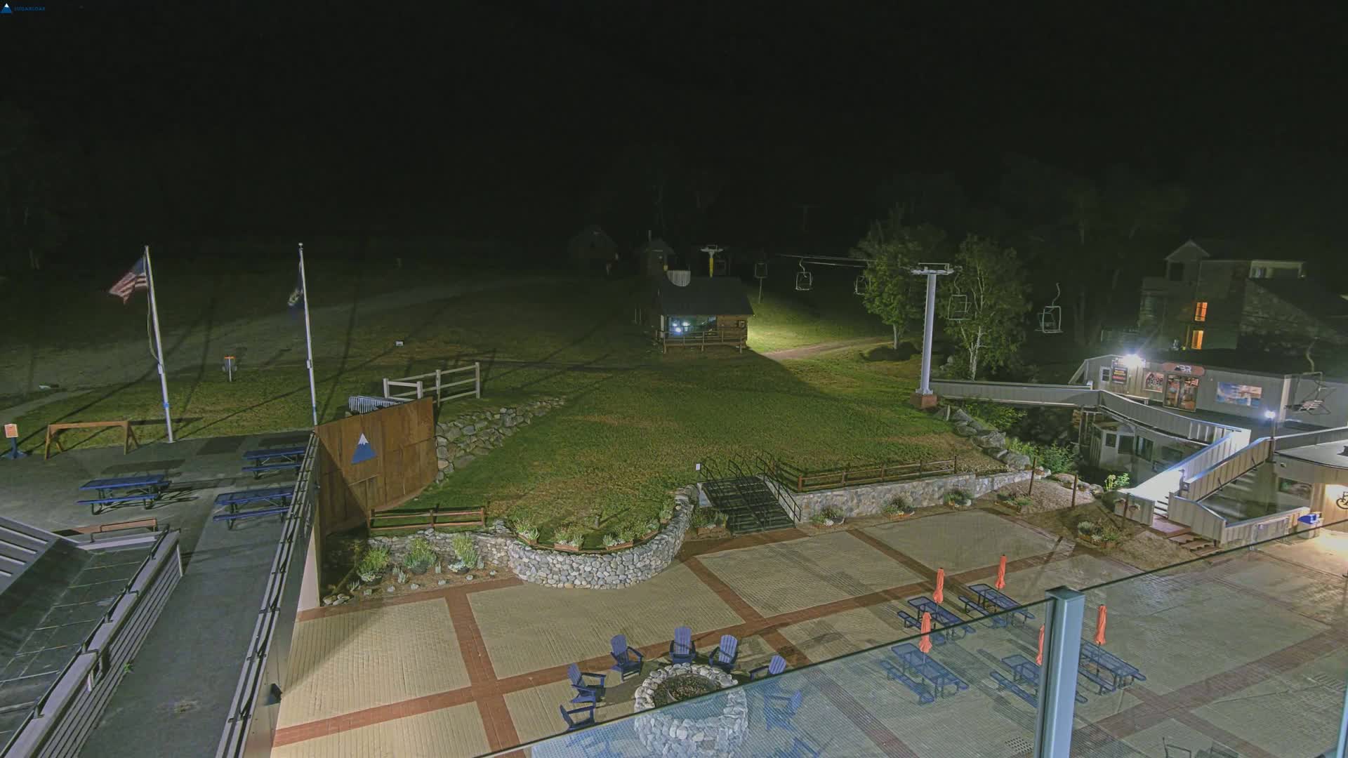 A nighttime view of a ski resort base area, featuring a paved patio with outdoor seating and fire pit, a small building, and a ski lift in the background under dark, clear skies.