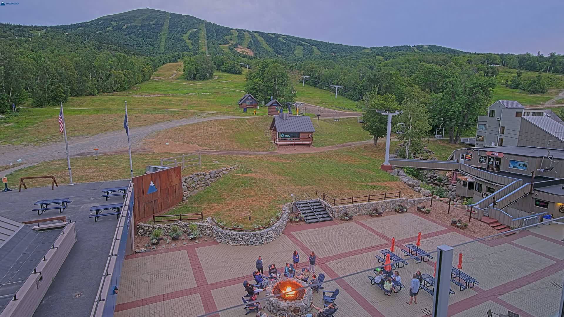 A paved patio area with picnic tables and a bonfire, surrounded by buildings and a grassy ski slope with chairlifts, under an overcast sky.