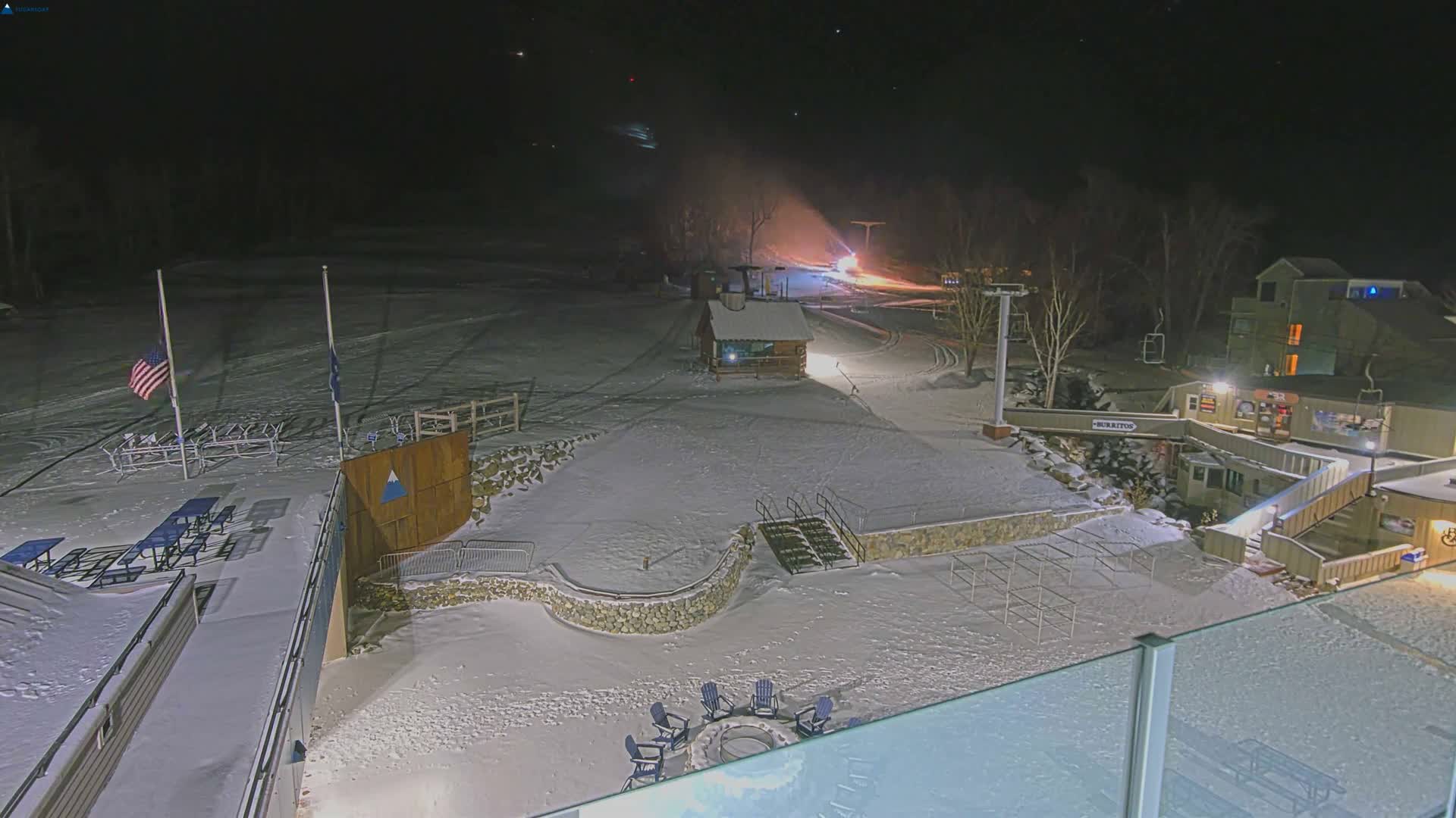 A clear and cold winter night illuminates a snow-covered ski resort, showcasing a working snow machine on a distant slope, a ski lift, various lodge buildings, and outdoor seating areas.