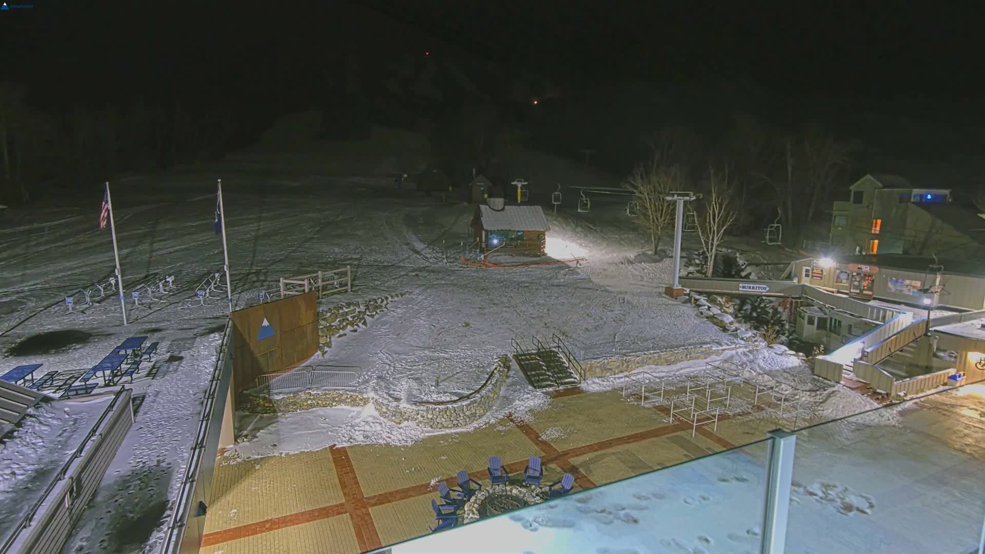A nighttime view of a snow-covered ski resort features illuminated buildings, dormant ski lifts, picnic areas, and a fire pit under a clear dark sky.