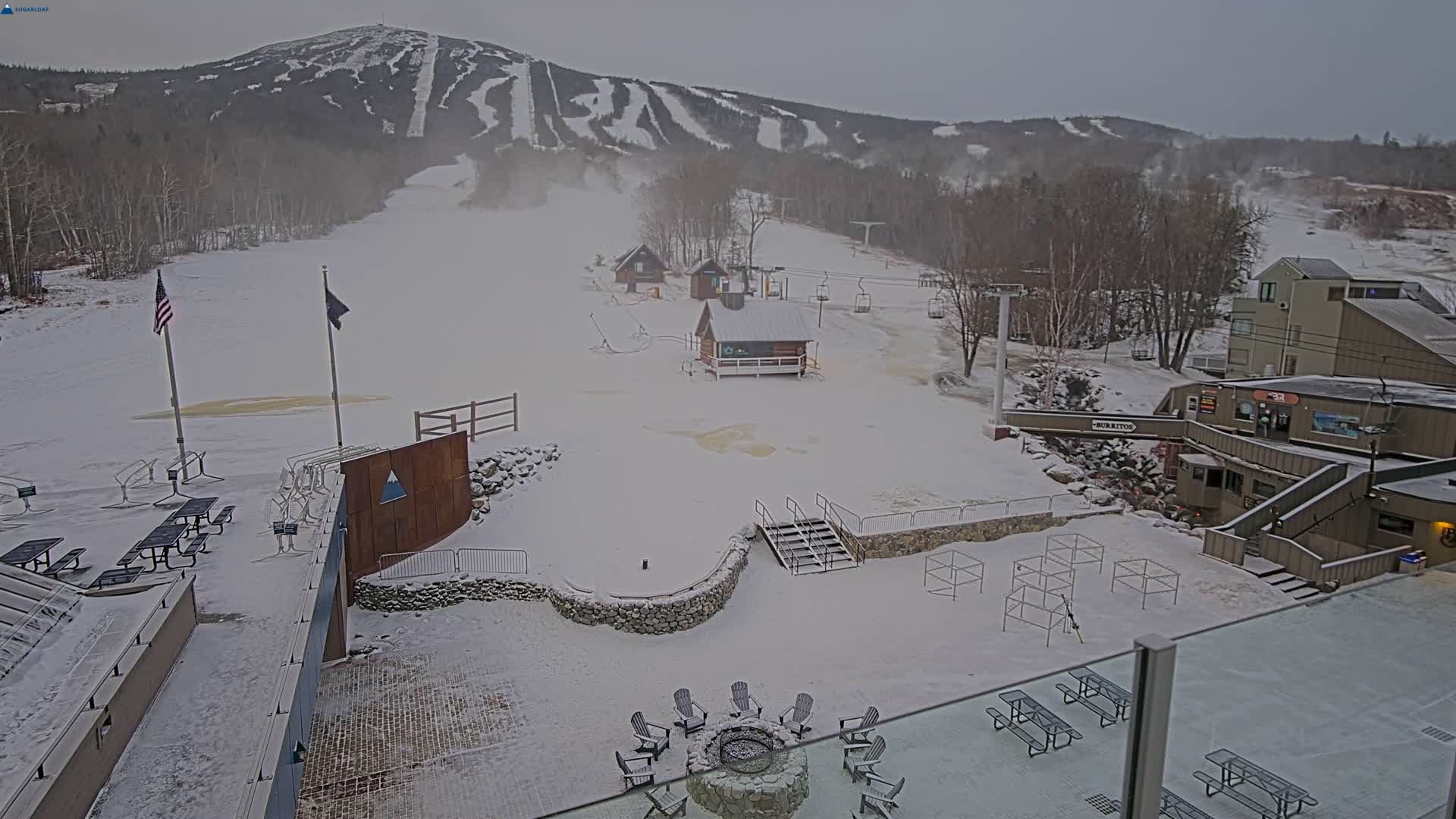 A nighttime view of a snow-covered ski resort features illuminated buildings, dormant ski lifts, picnic areas, and a fire pit under a clear dark sky.