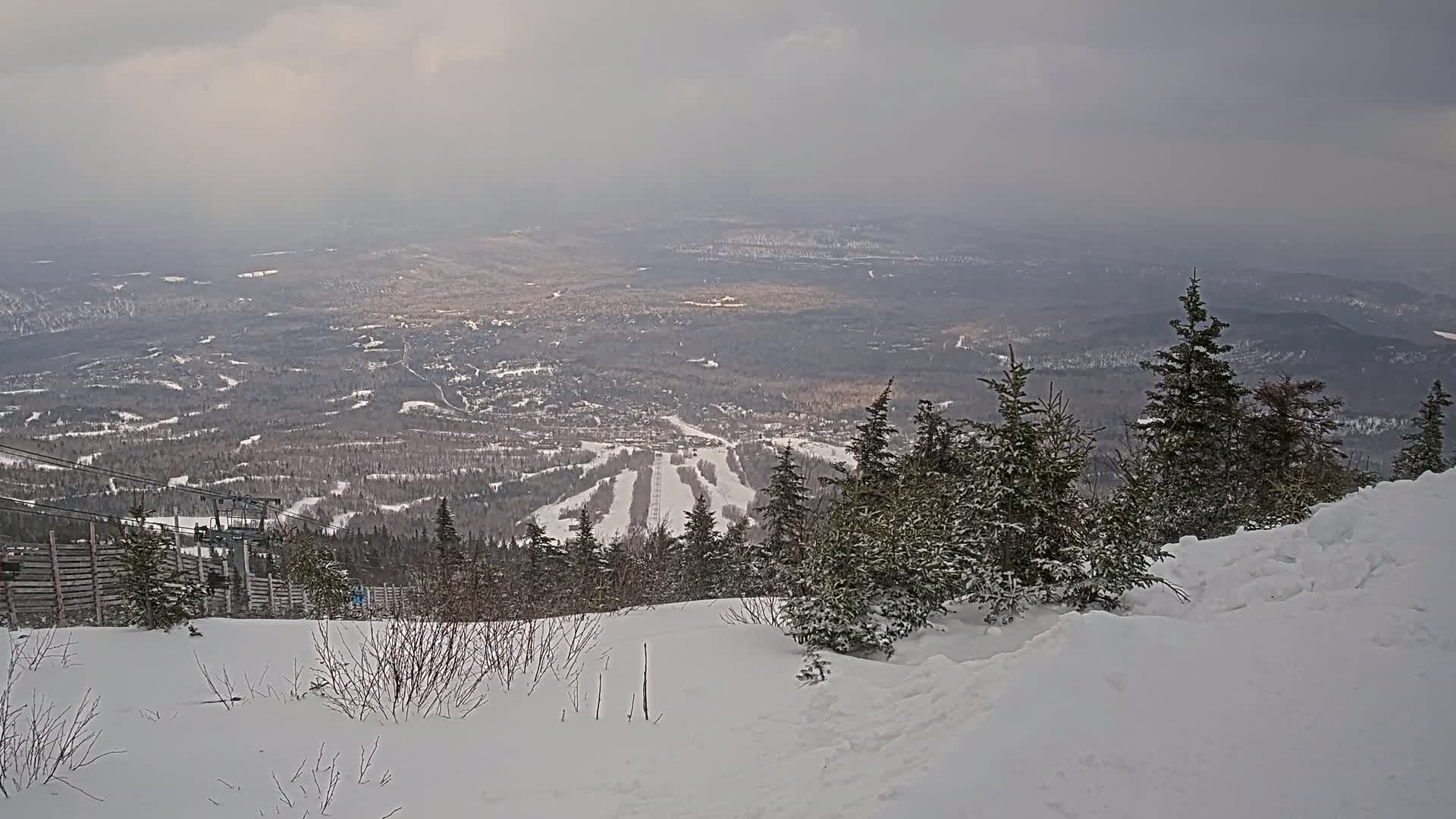 A snowy, overcast mountaintop view reveals a ski resort valley below with a chairlift and snow-covered evergreen trees in the foreground.