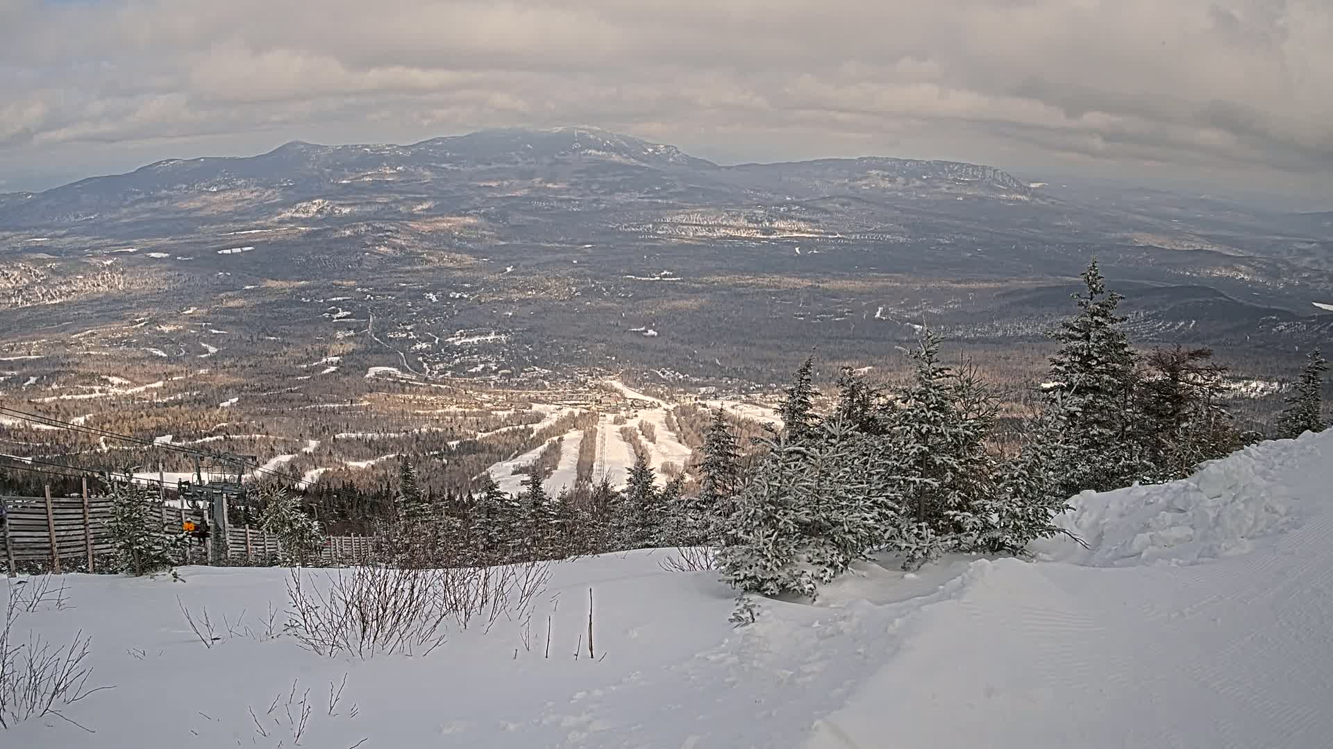 A snowy mountaintop view reveals a ski resort nestled in a valley below, under a cloudy sky.