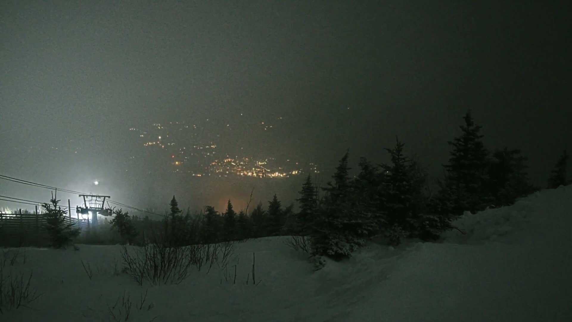 A snow-covered landscape at night, dimly lit by a distant town and a ski lift, is shrouded in fog.