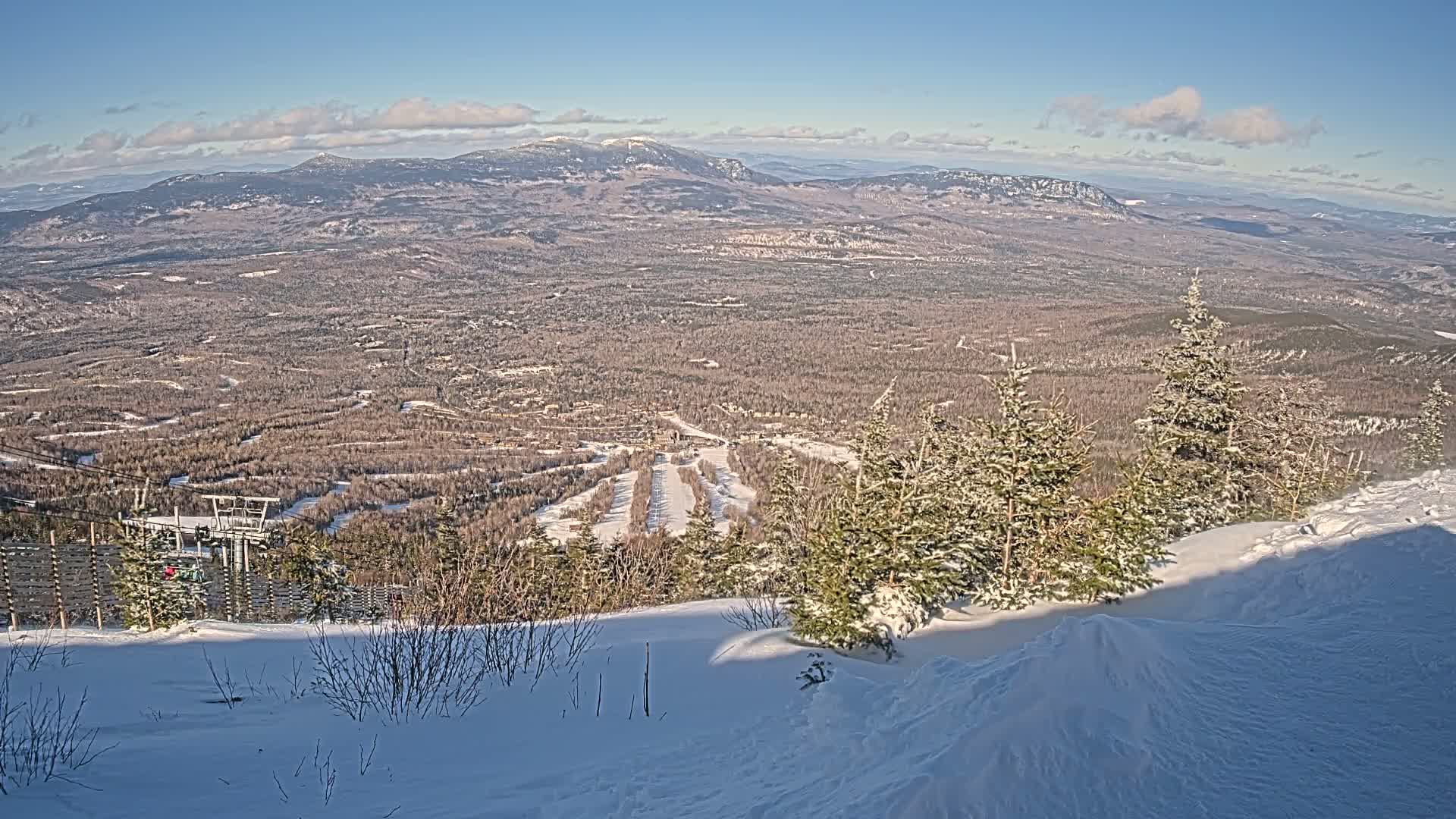 A snow-covered mountaintop offers a panoramic view of a vast, snow-dusted forest and distant mountains under a mostly clear, sunny sky.