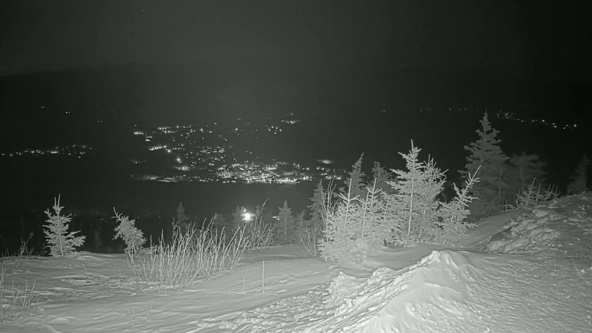 A snow-covered hill at night overlooks a distant town illuminated by lights.