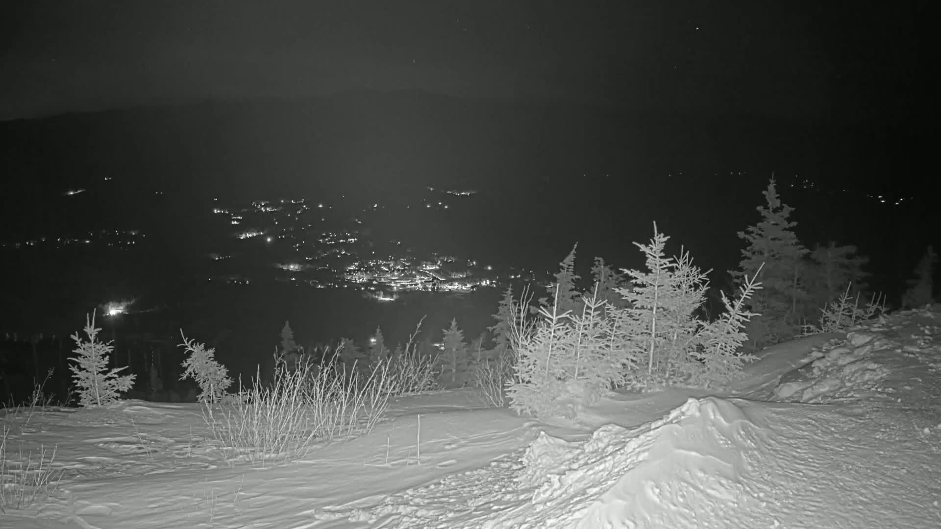 A snowy, nighttime landscape shows a valley town illuminated below a snow-covered hill dotted with evergreen trees.