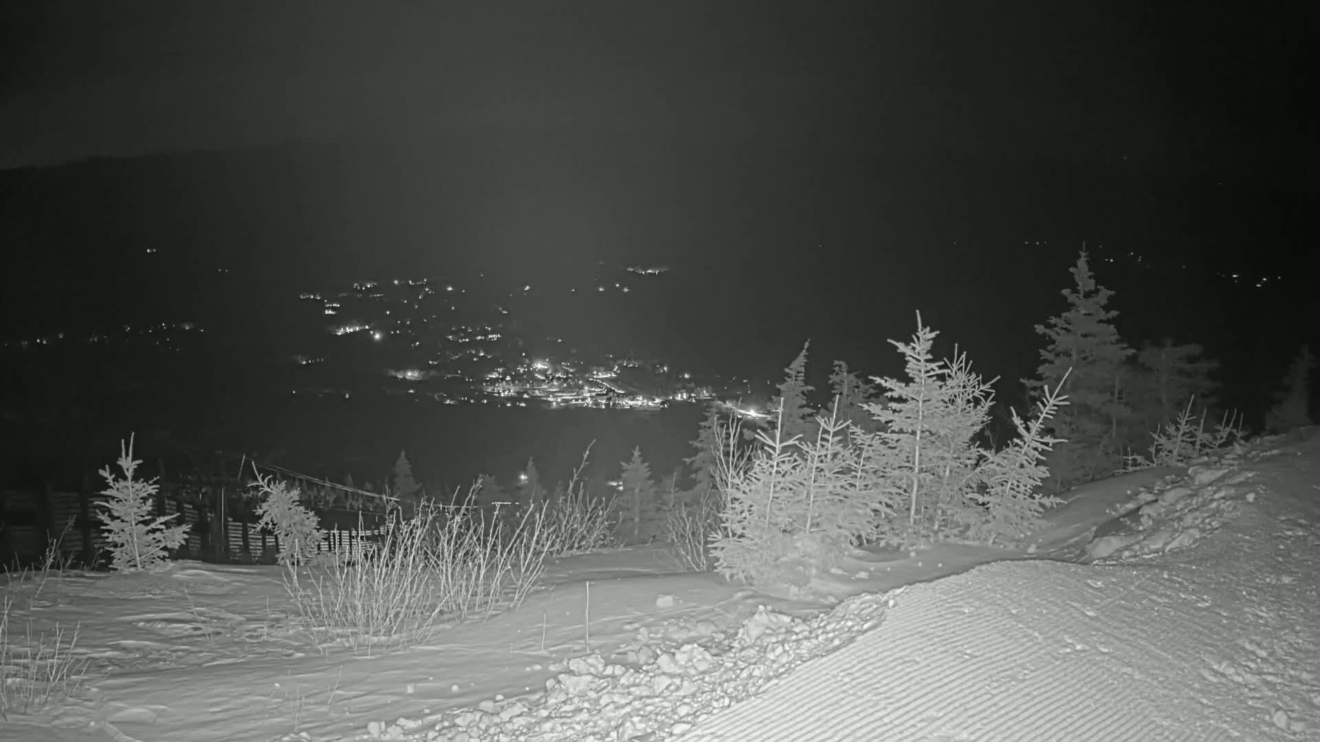A snow-covered hillside at night overlooks a distant town illuminated by lights.