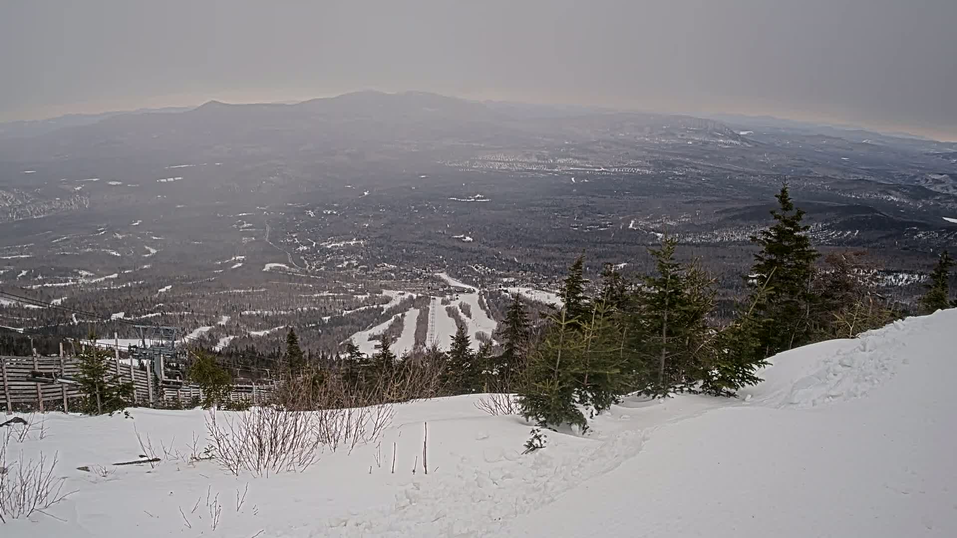 A snowy mountain slope overlooks a vast, hazy valley with a ski resort visible in the distance under an overcast sky.