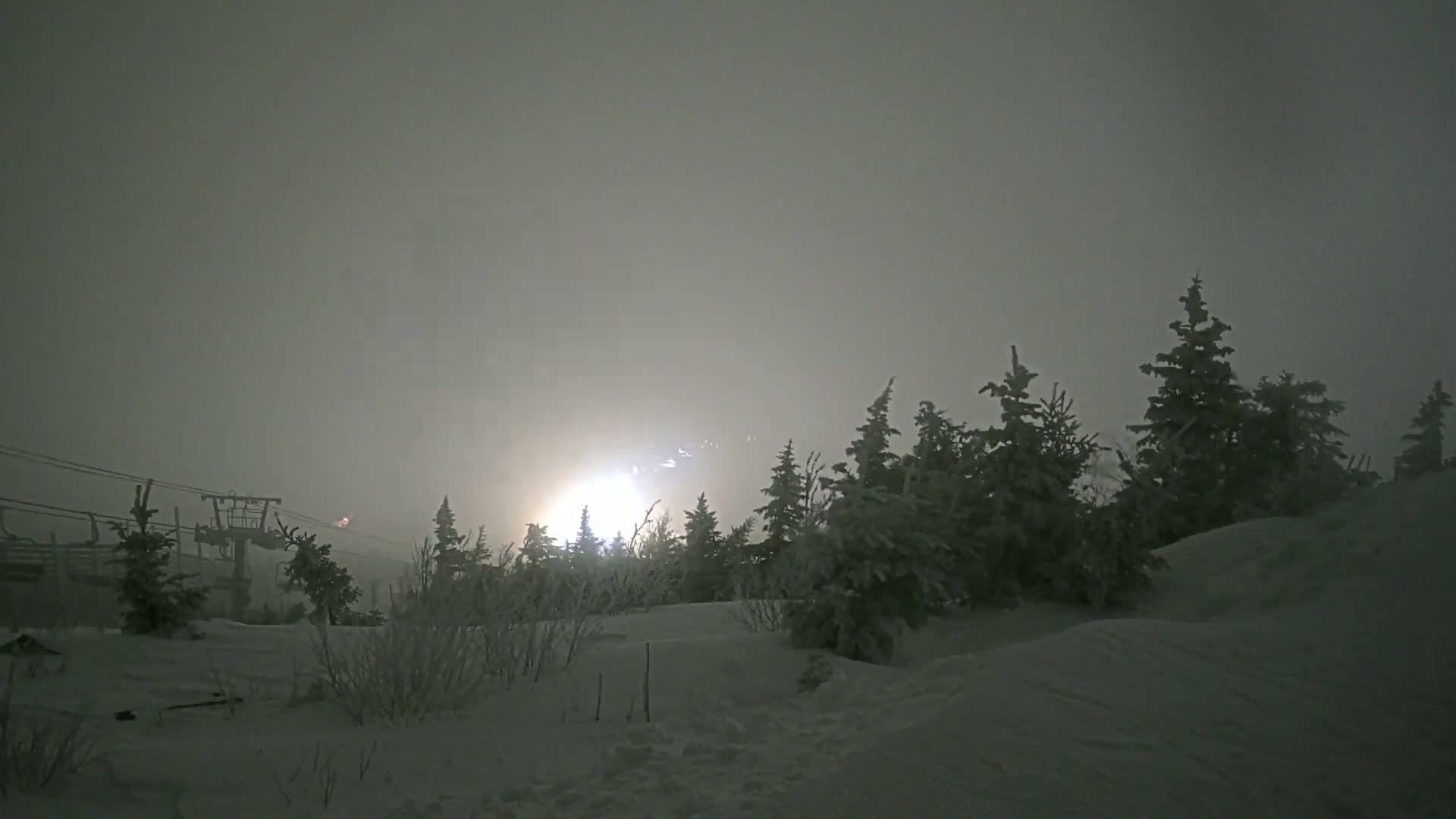 A snowy, foggy landscape at night features a ski lift in the distance illuminated by lights, with snow-covered evergreen trees in the foreground.