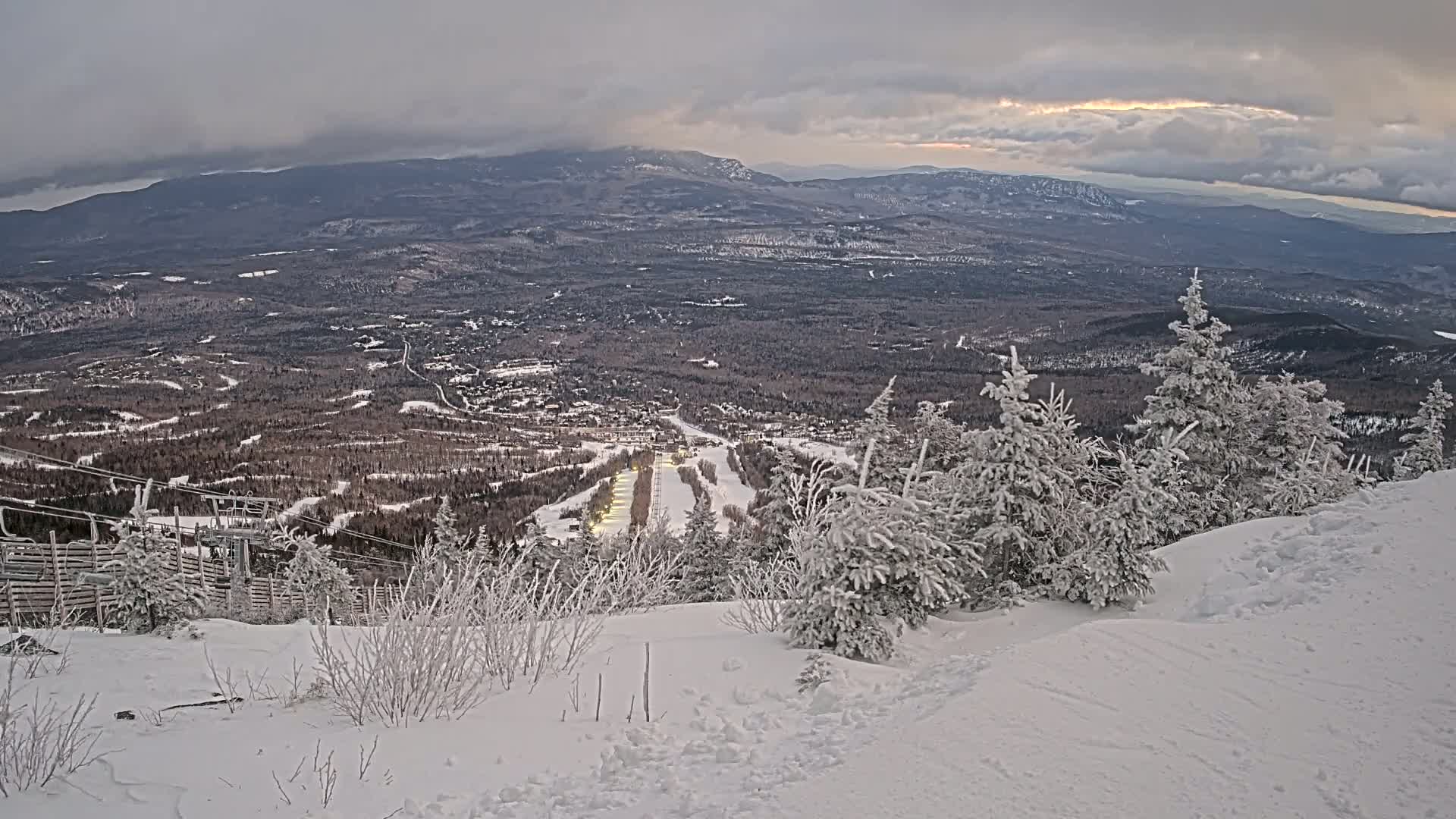 A snow-covered mountain slope overlooks a valley with a ski resort, under a cloudy but clearing sky.
