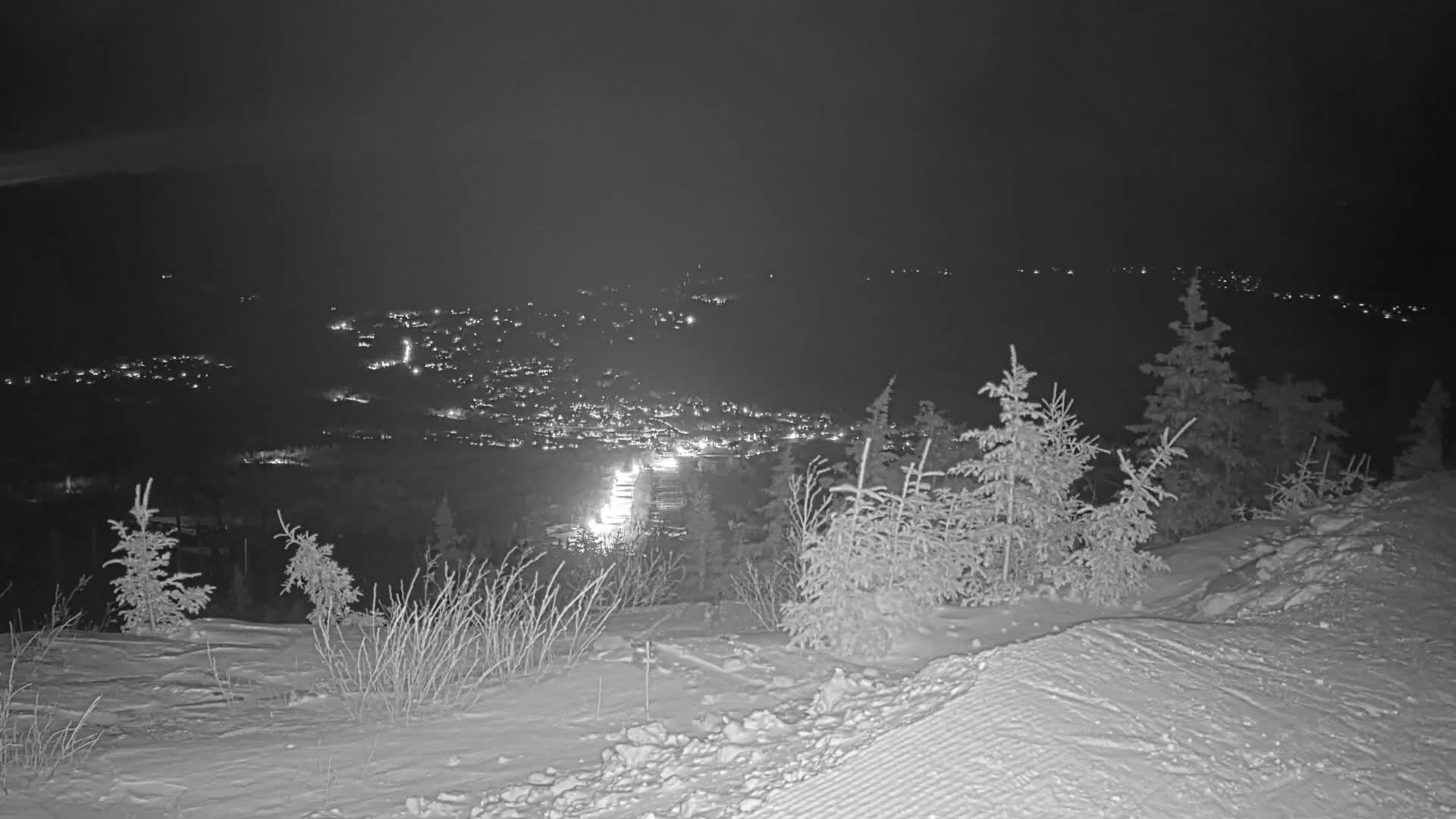 A snowy, nighttime landscape shows a distant town illuminated by lights, viewed from a snow-covered hilltop with small evergreens.
