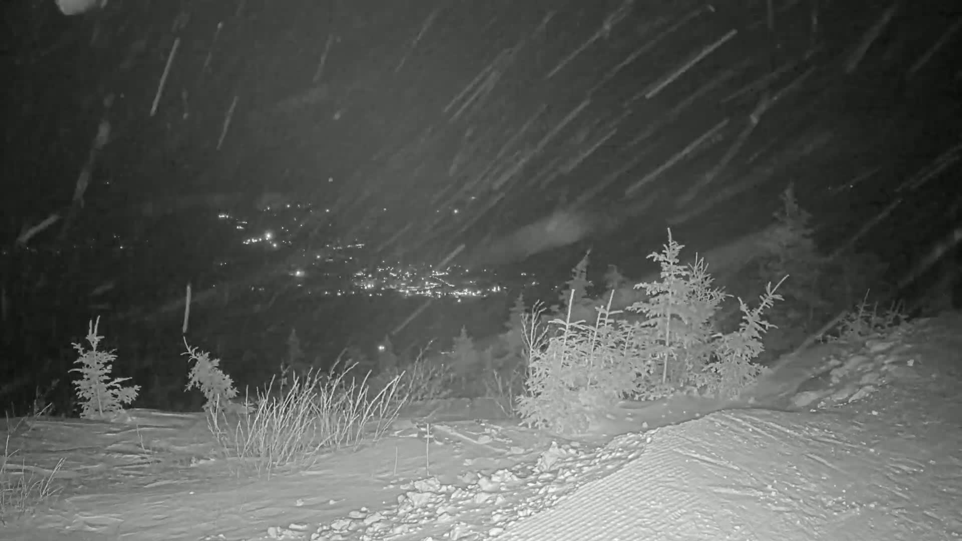 A snowy nighttime scene shows a distant town illuminated below a snow-covered hill with small evergreen trees.