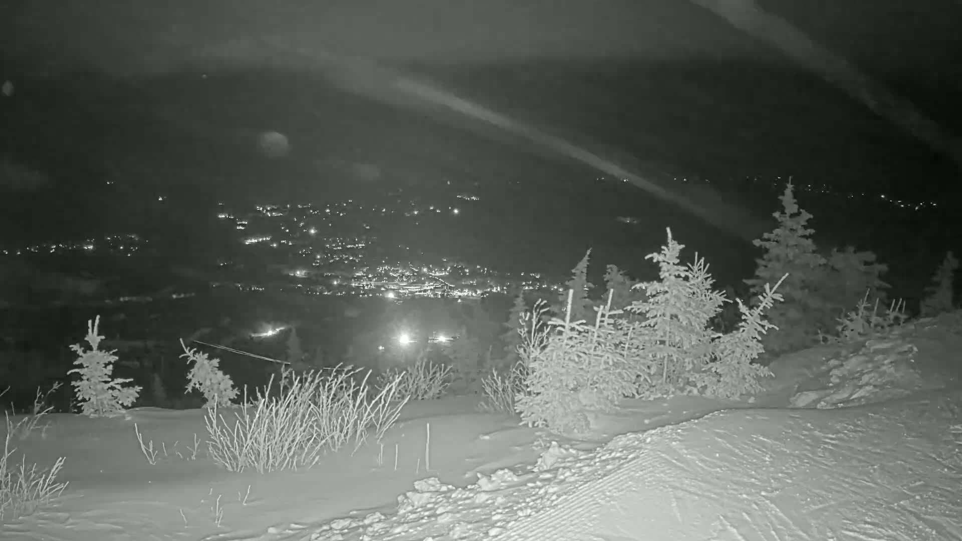 A nighttime, snowy landscape shows a distant town illuminated below a snow-covered hill with small evergreen trees.