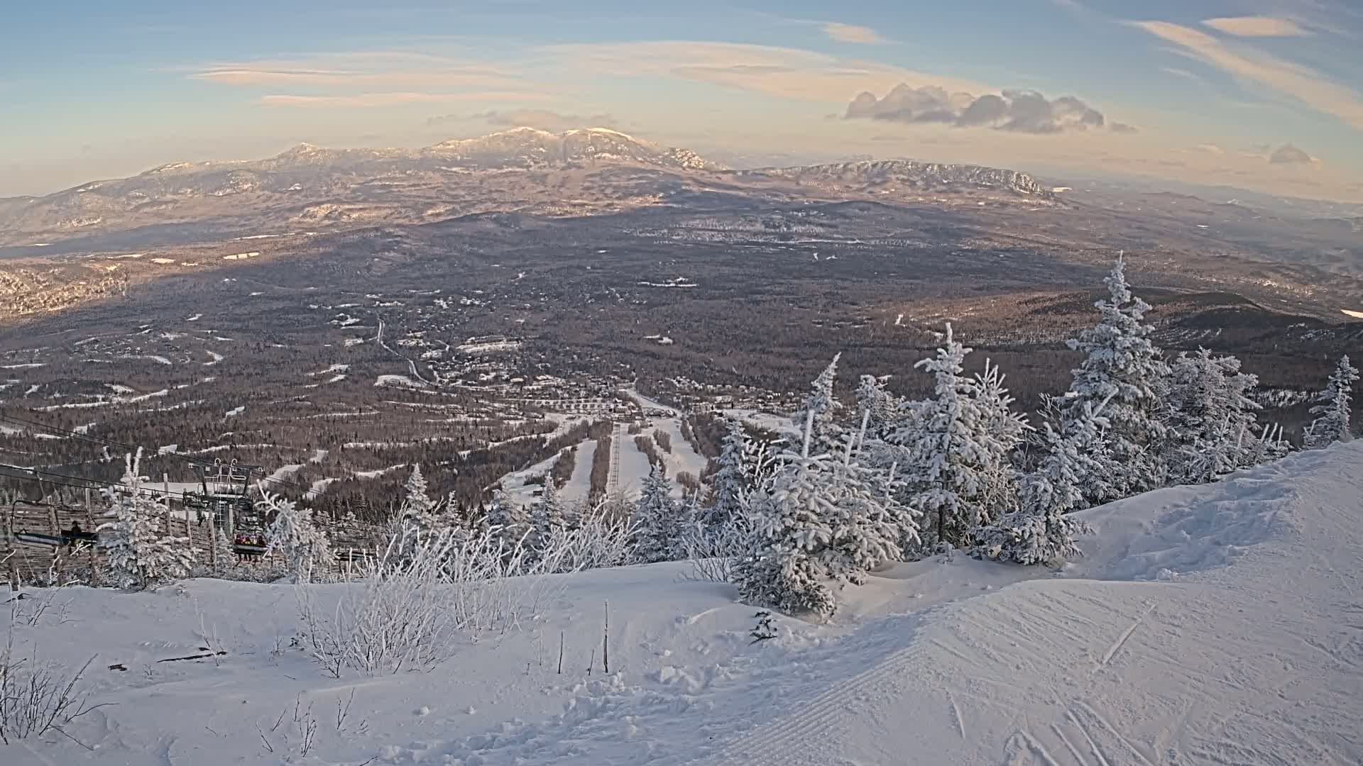 A snowy mountaintop view reveals a ski resort nestled in a valley below, under a partly cloudy, bright sky.