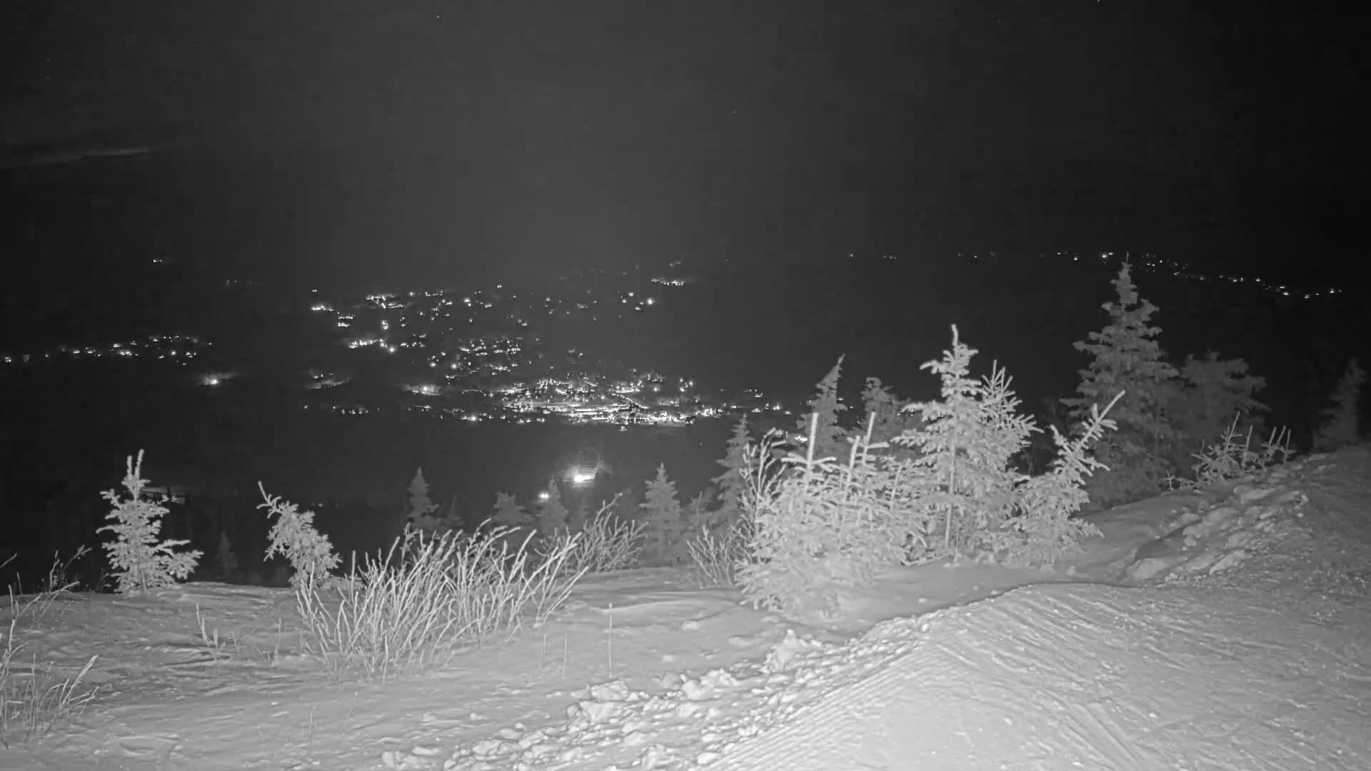 A nighttime, snowy landscape shows a distant town illuminated below a snow-covered hill with evergreen trees.