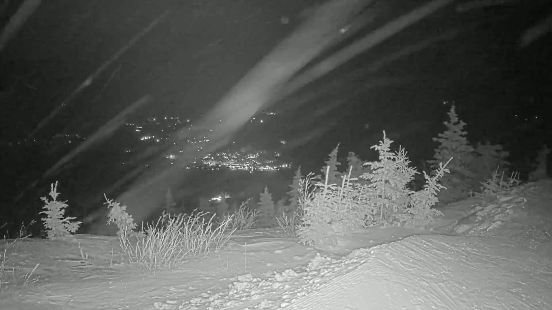 A snowy, nighttime scene shows a distant town's lights below a snow-covered hillside with several small evergreen trees.