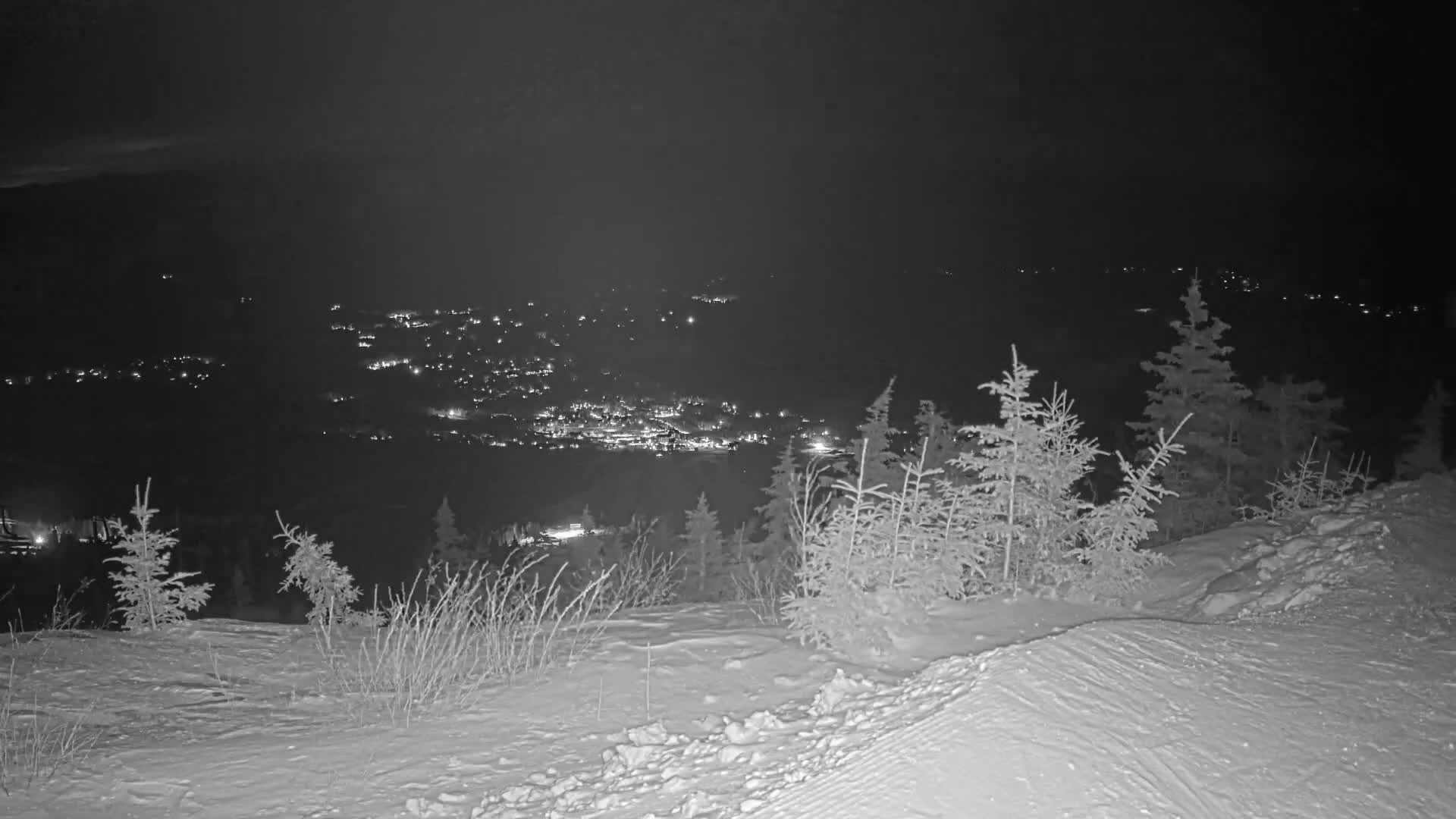 A snowy, nighttime hillside overlooks a distant town illuminated by lights.