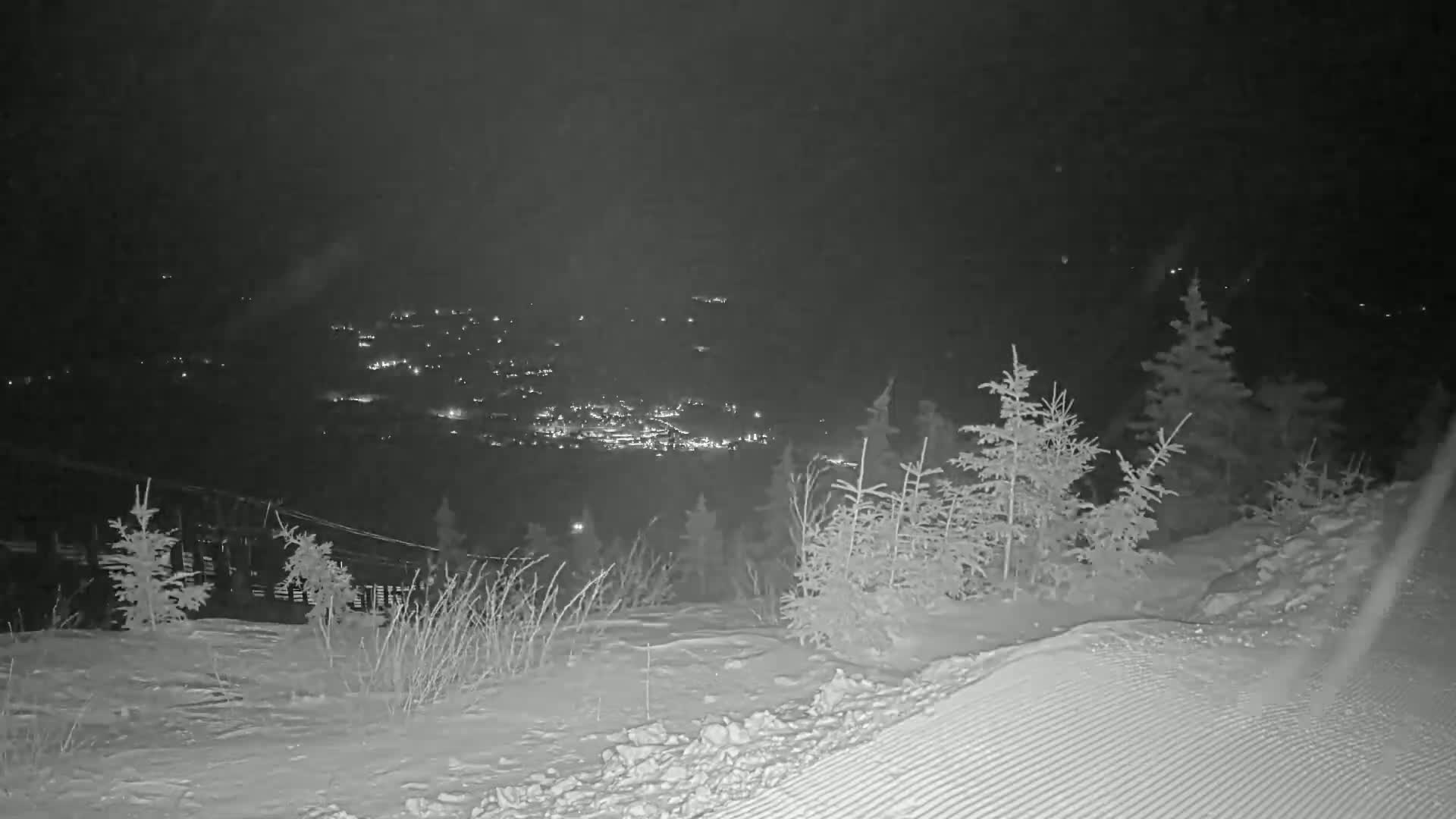 A nighttime view from a snowy hilltop shows a distant town illuminated below, under a dark, possibly overcast sky.
