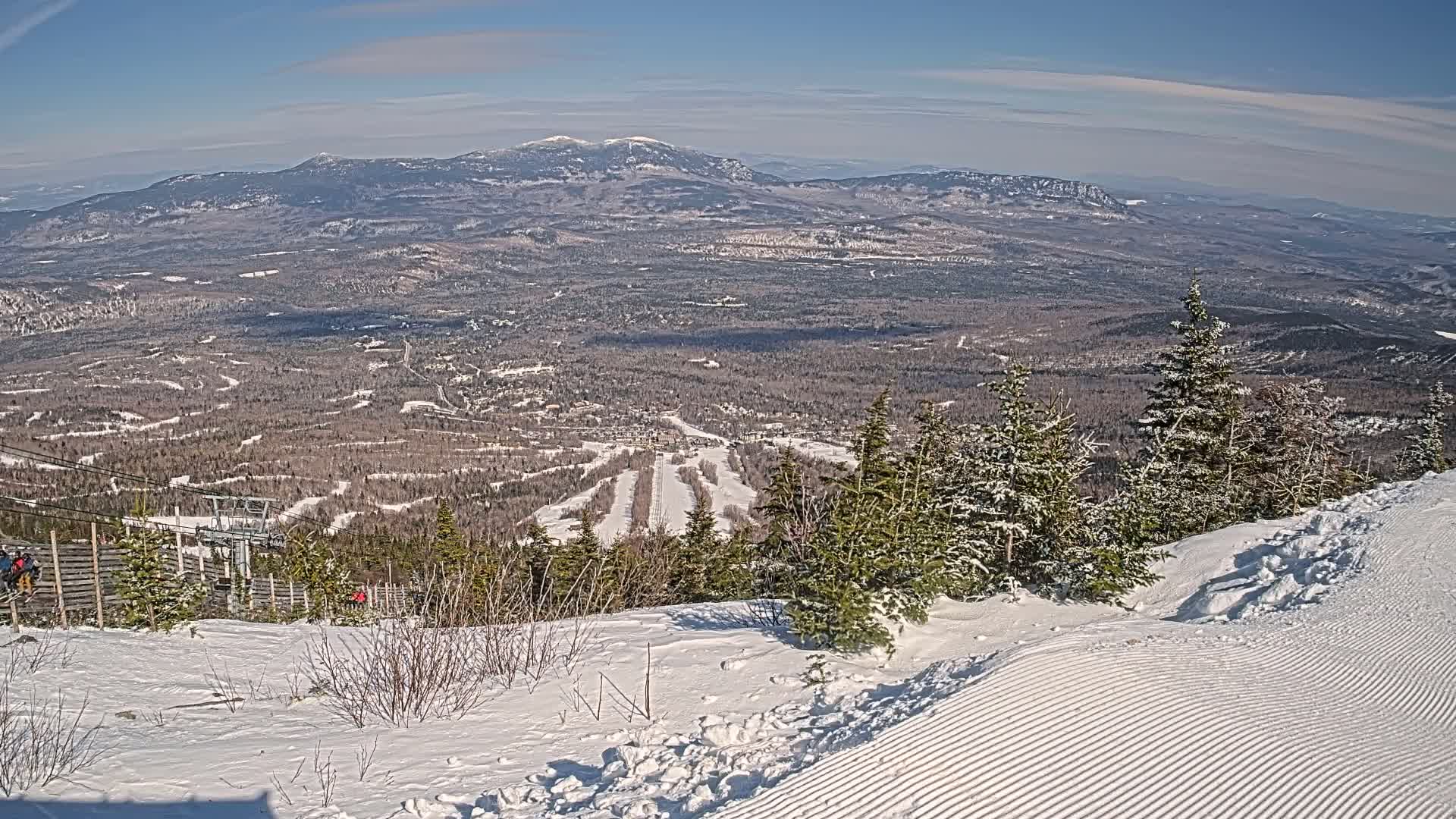A snow-covered mountaintop overlooks a vast, snow-dusted forest and distant mountains under a clear blue sky.