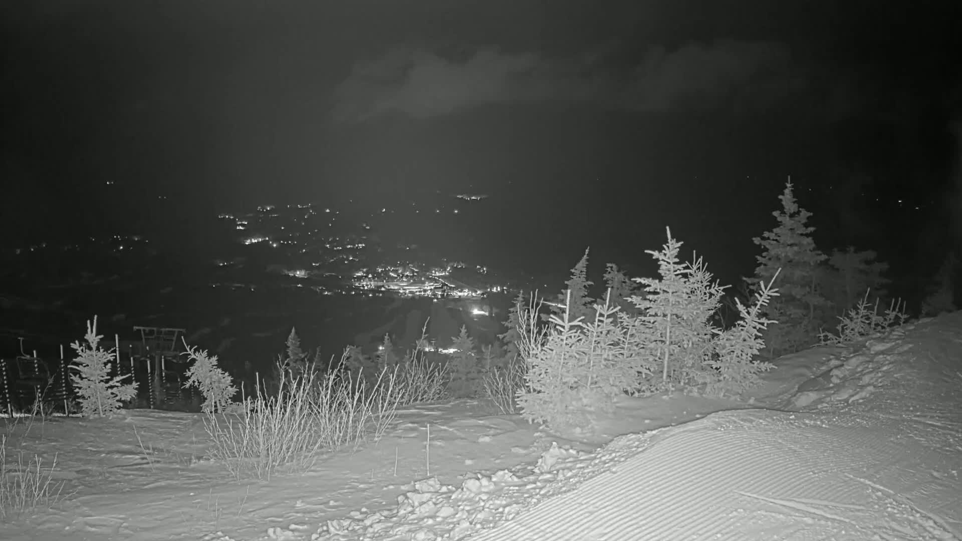 A snowy, nighttime landscape shows a valley with distant lights below a snow-covered hill dotted with evergreen trees under a cloudy sky.