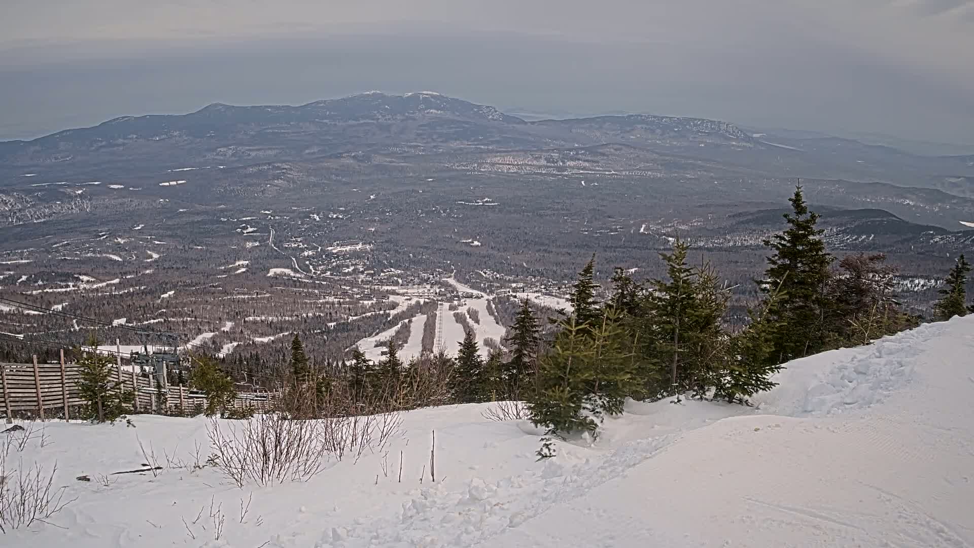 A snow-covered mountain slope overlooks a valley with a ski resort and distant mountains under an overcast sky.