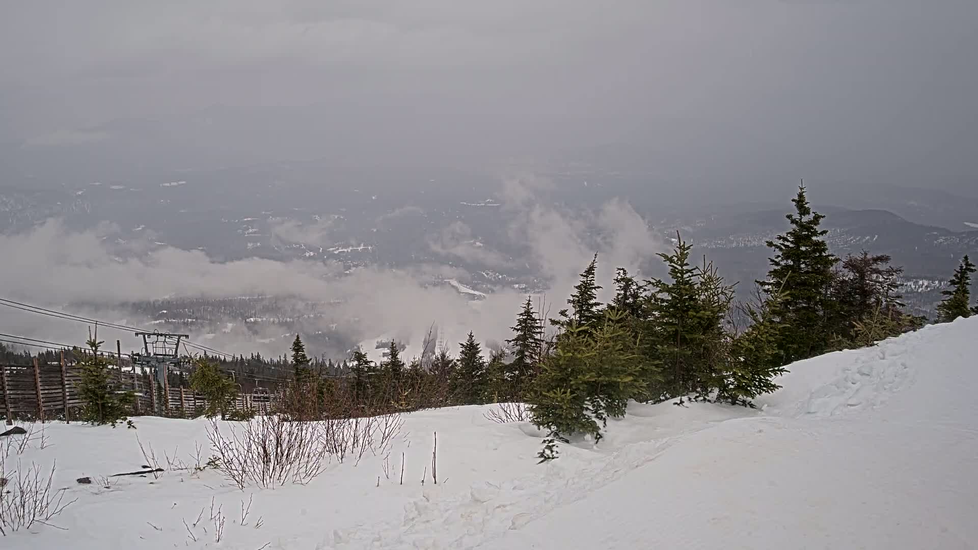 A snowy mountaintop view shows a ski lift and evergreen trees in the foreground, with a valley shrouded in fog below on an overcast day.