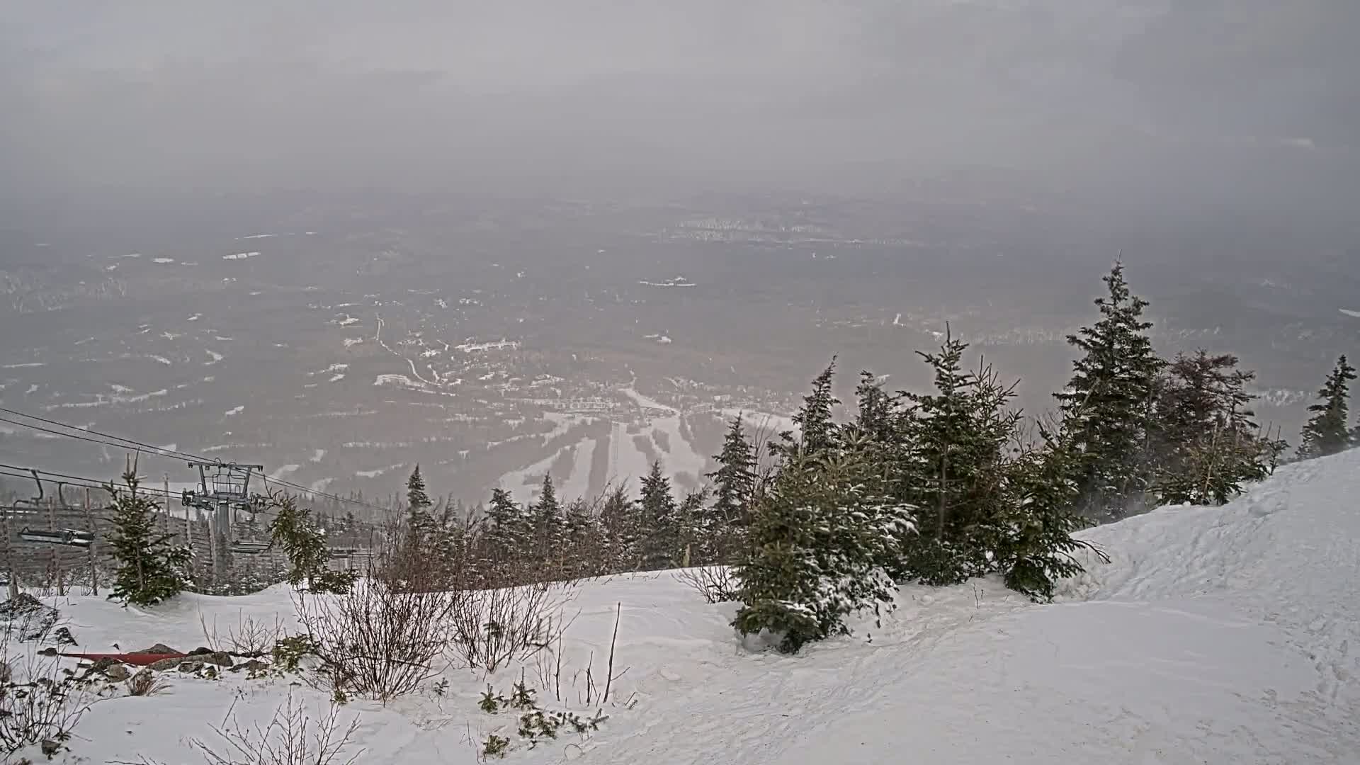 A snow-covered ski resort is visible in the distance, with snow-covered evergreens in the foreground, under hazy, overcast conditions.