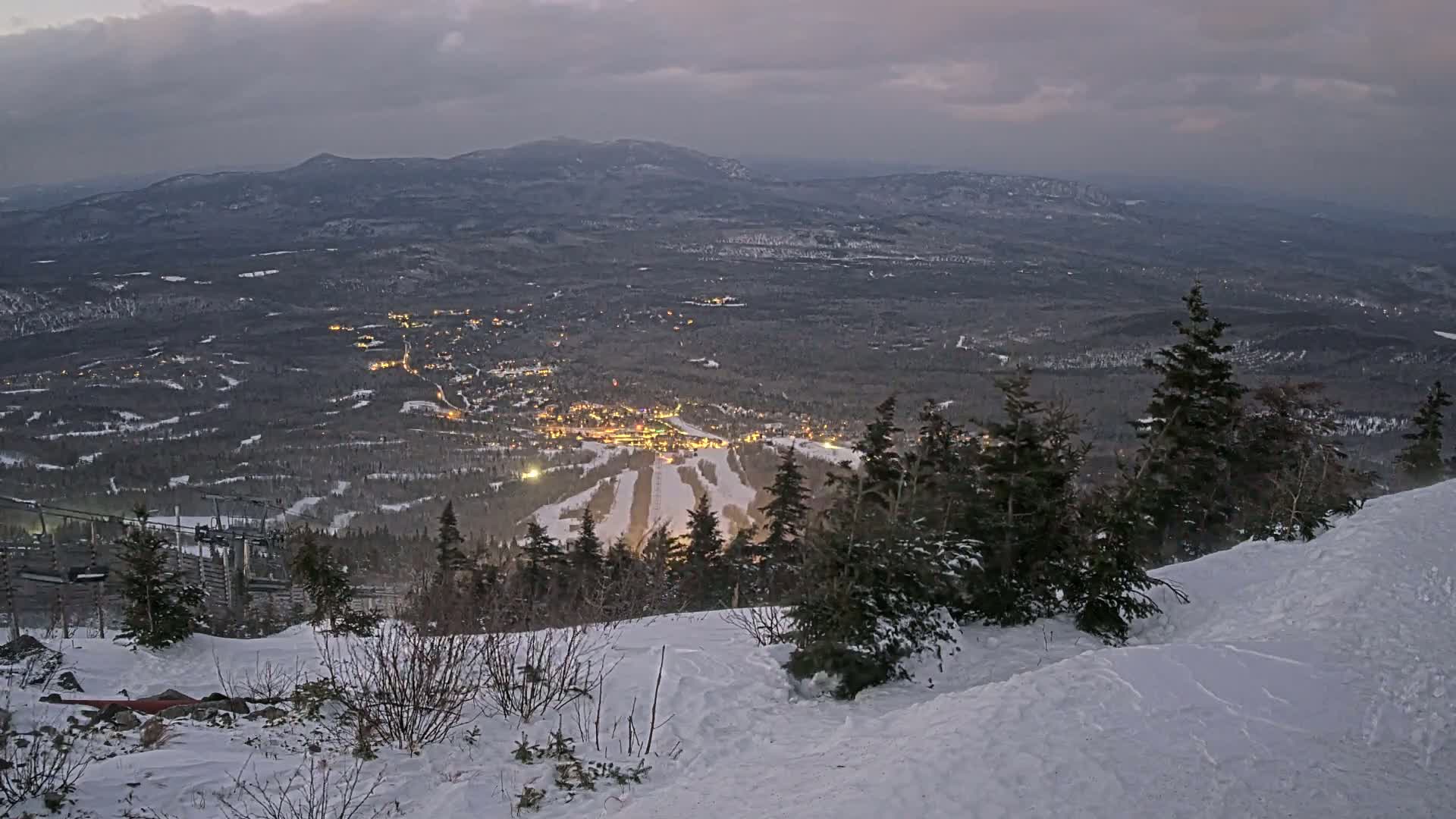 A snow-covered mountain slope overlooks a valley town illuminated at twilight under a cloudy sky.