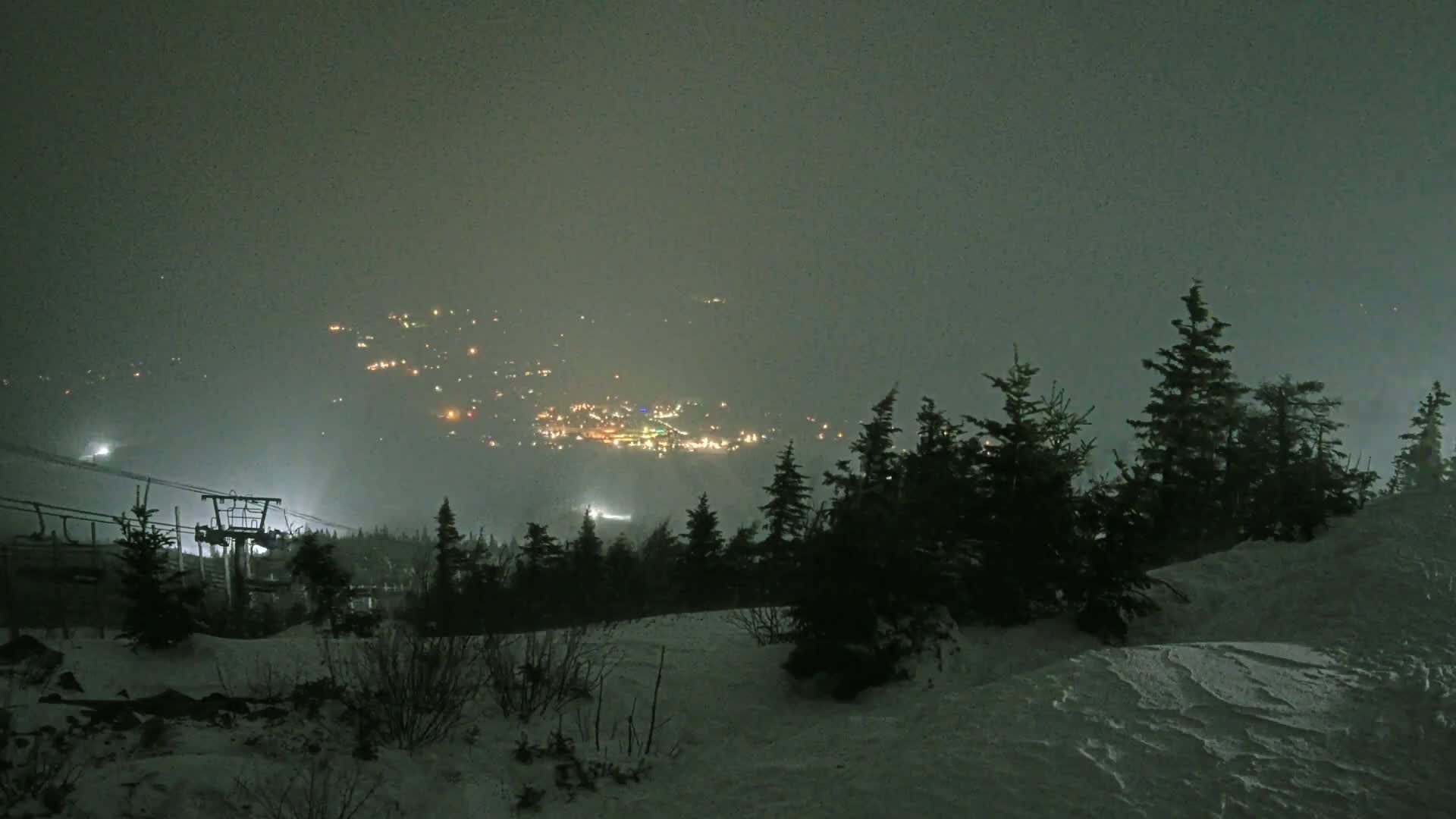 A snow-covered ski resort at night, shrouded in fog, with a chairlift visible in the foreground.