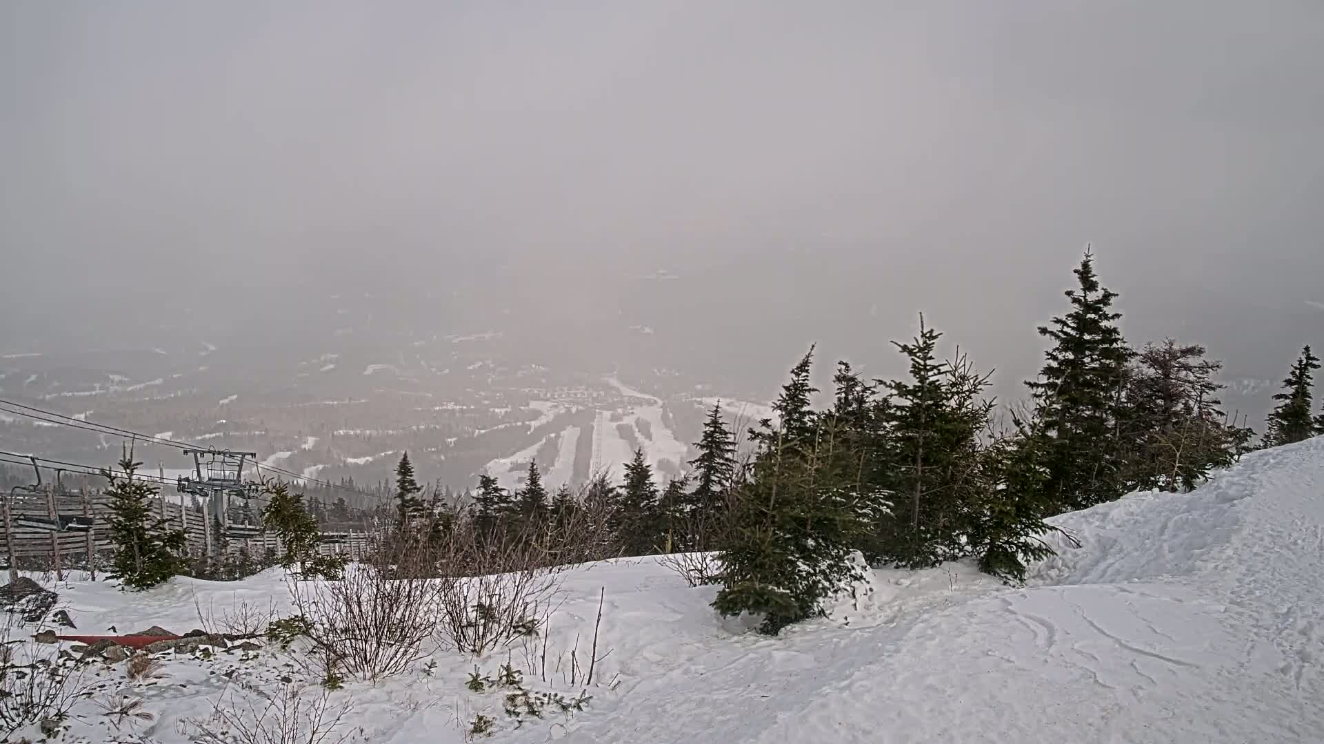 A snow-covered ski resort is viewed from a snowy mountaintop on an overcast day, with a chairlift visible in the foreground.