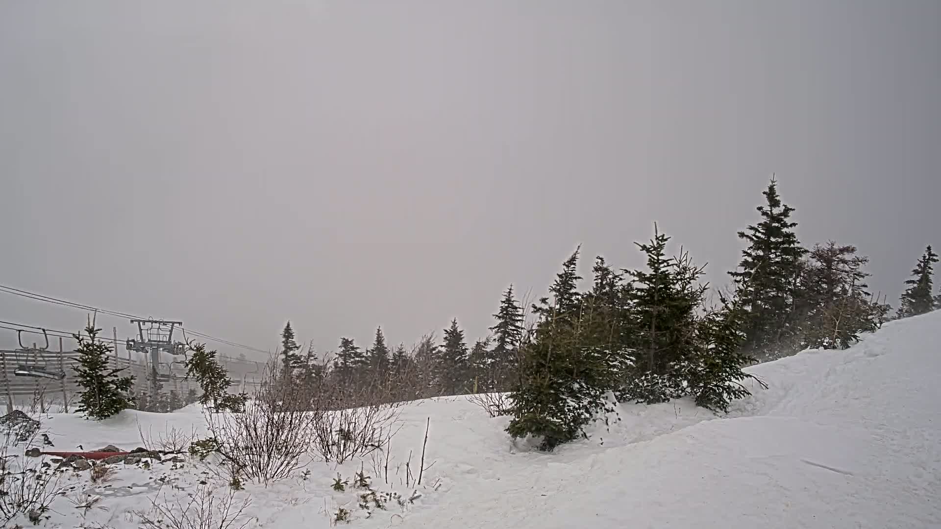 A snowy, overcast landscape features a ski lift and evergreen trees partially obscured by blowing snow.