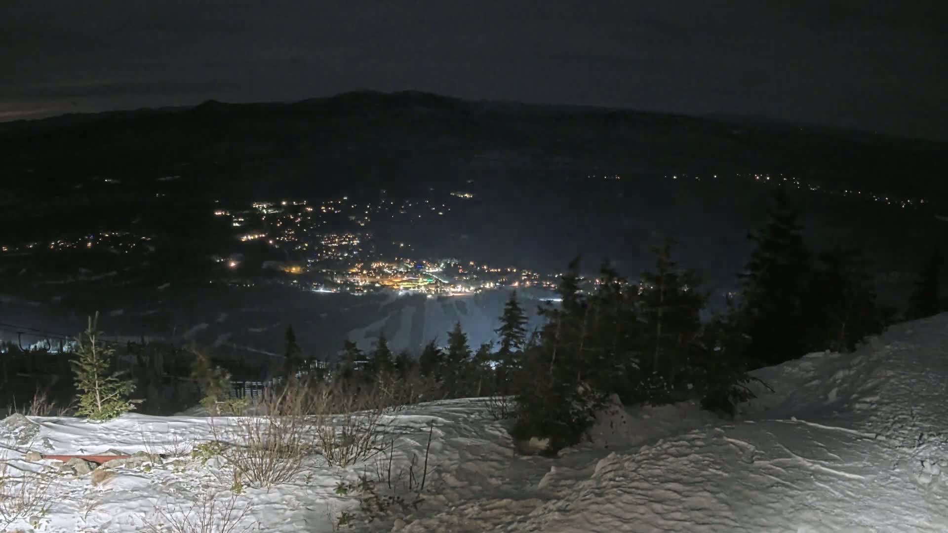 A snow-covered hillside at night overlooks a valley town illuminated by lights, under a dark sky.