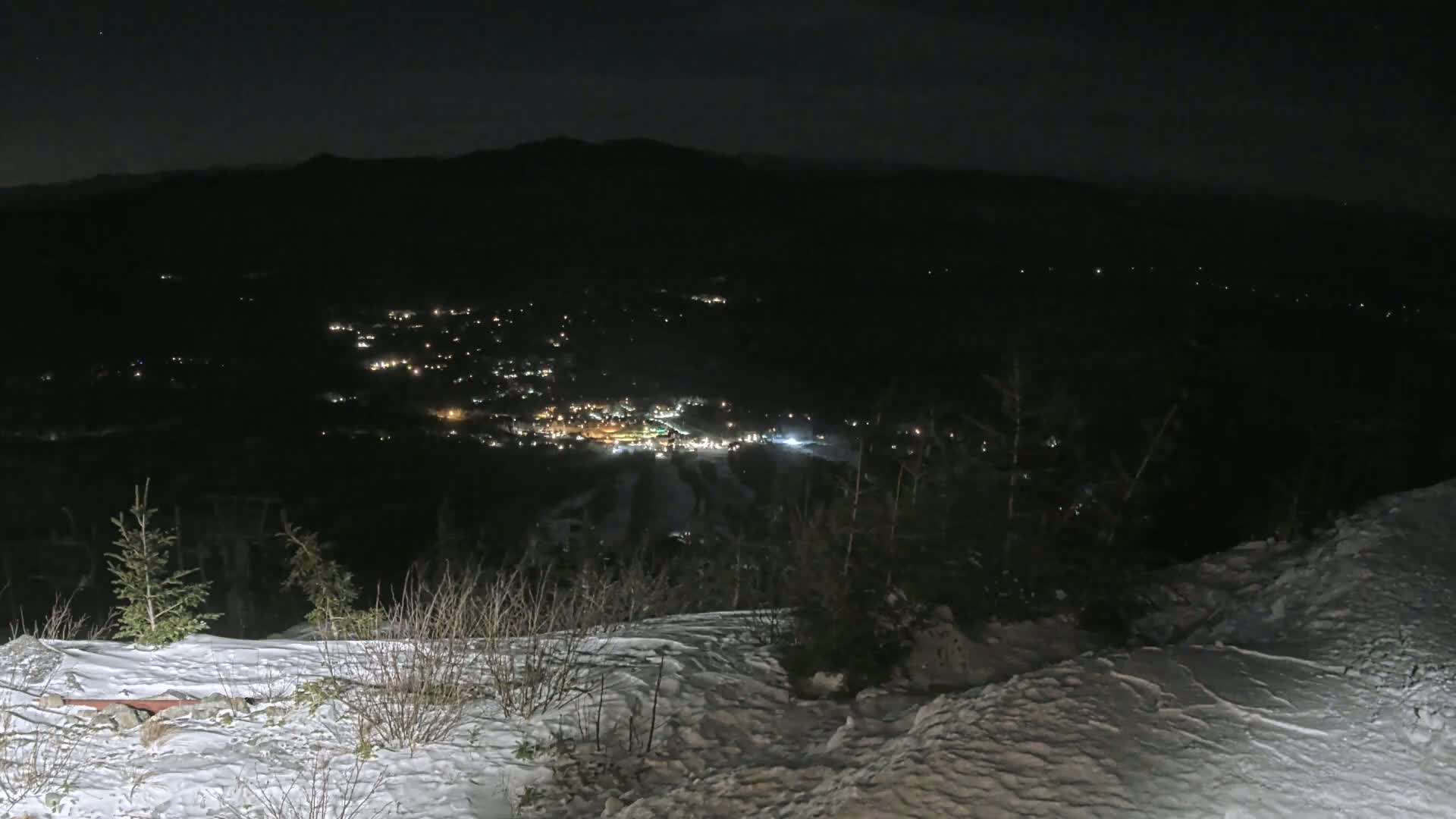 A snow-covered hillside at night overlooks a valley illuminated by the lights of a town below.