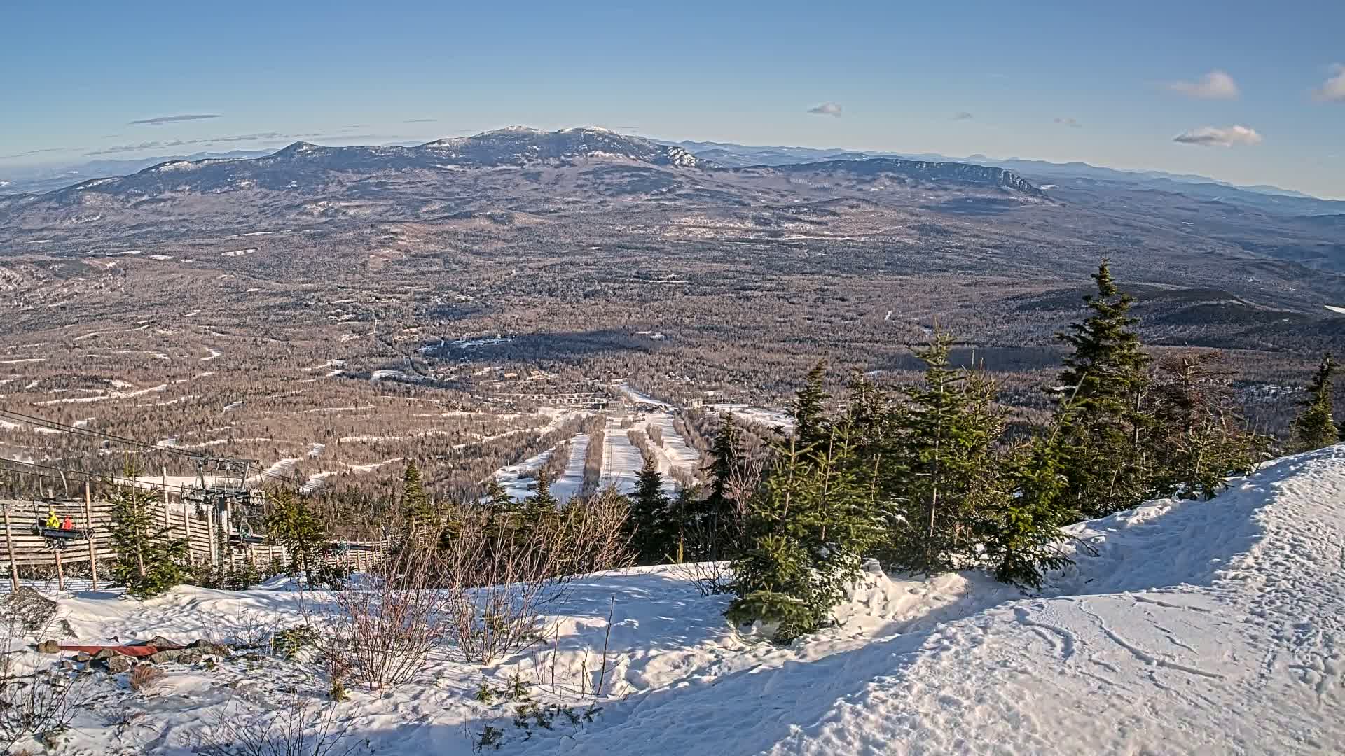 A snowy mountaintop view overlooking a vast, snow-dusted forest and distant mountains under a clear blue sky.