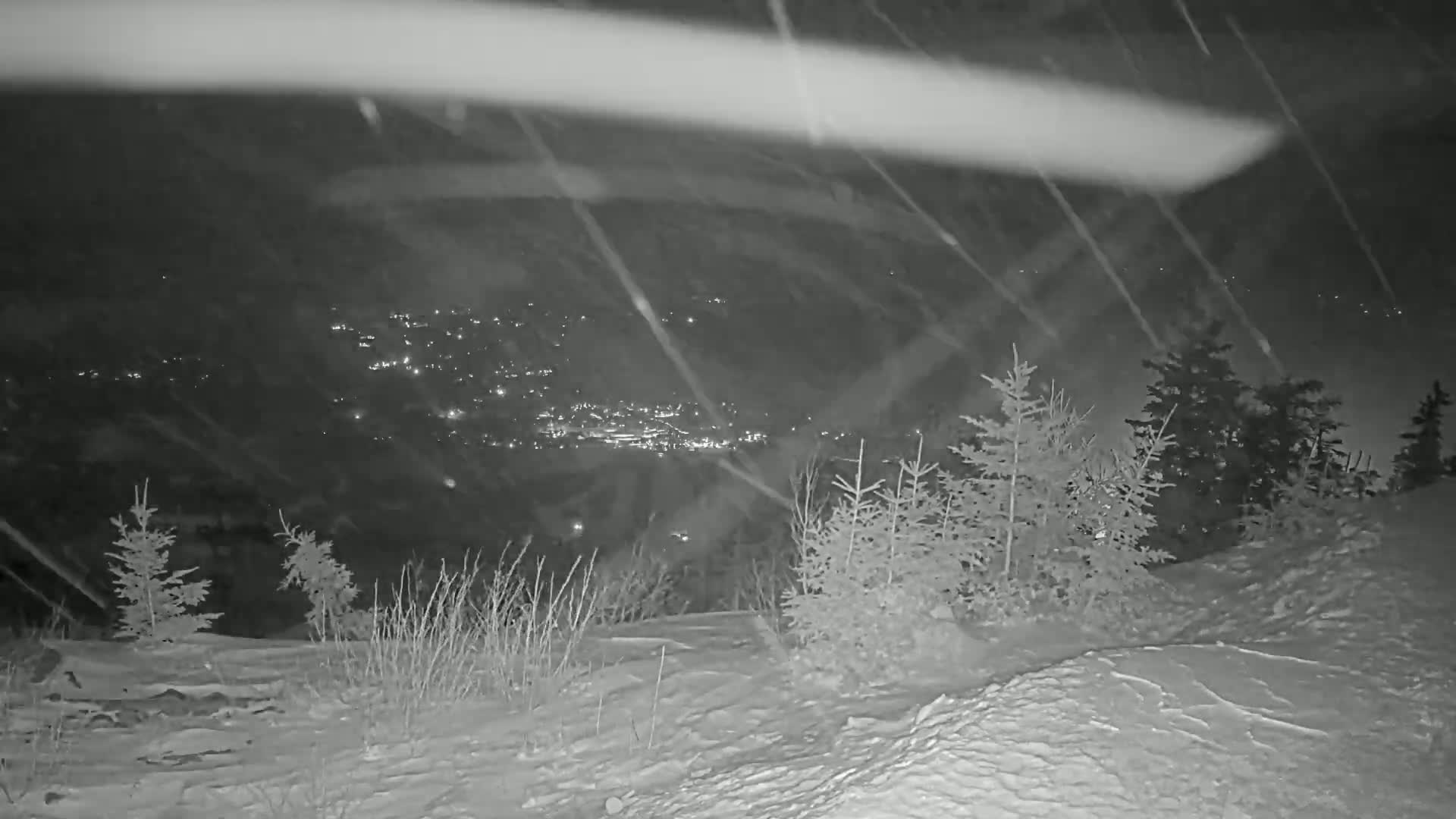 A snowy, nighttime view shows a distant town's lights below a snow-covered hillside with evergreen trees in the foreground, under heavy snowfall.