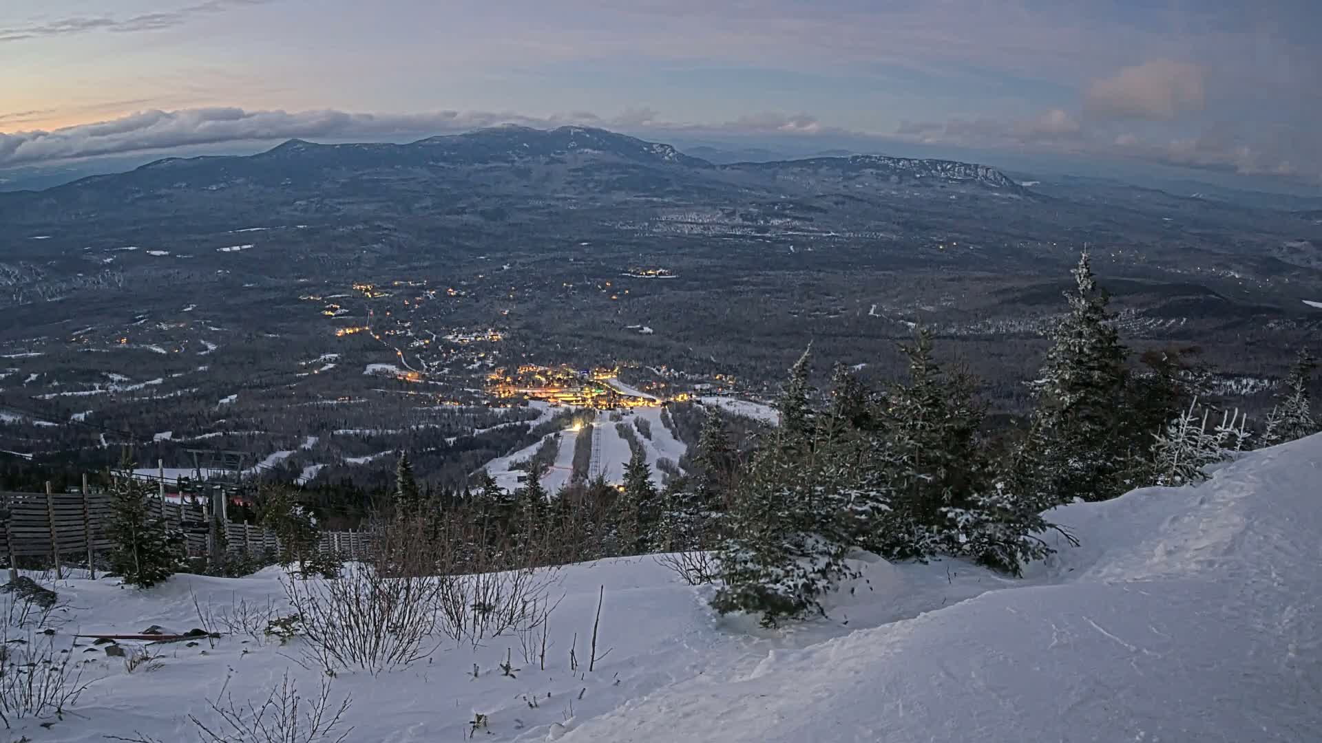 A snow-covered mountain slope overlooks a valley with a lit-up town and ski resort at twilight under a cloudy sky.