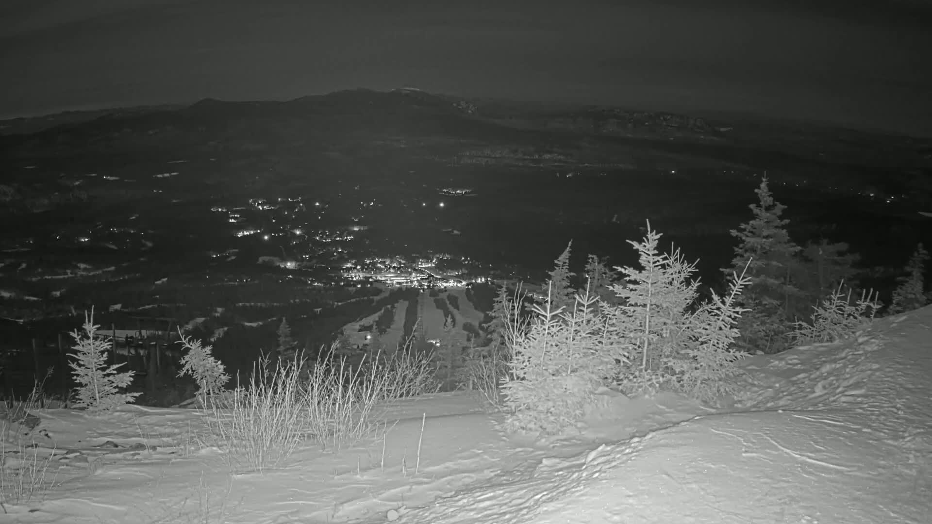 A nighttime, snowy mountaintop view overlooks a valley town illuminated by lights, with dark mountains in the background.
