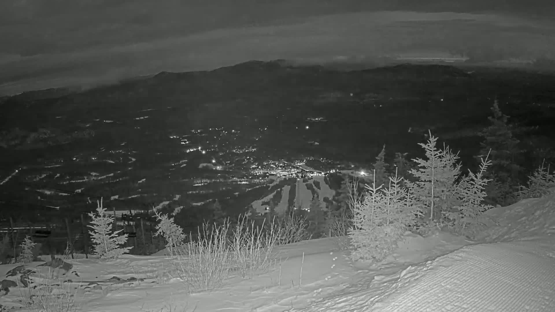 A snow-covered hillside at night overlooks a valley with a town and ski resort, under a cloudy sky.