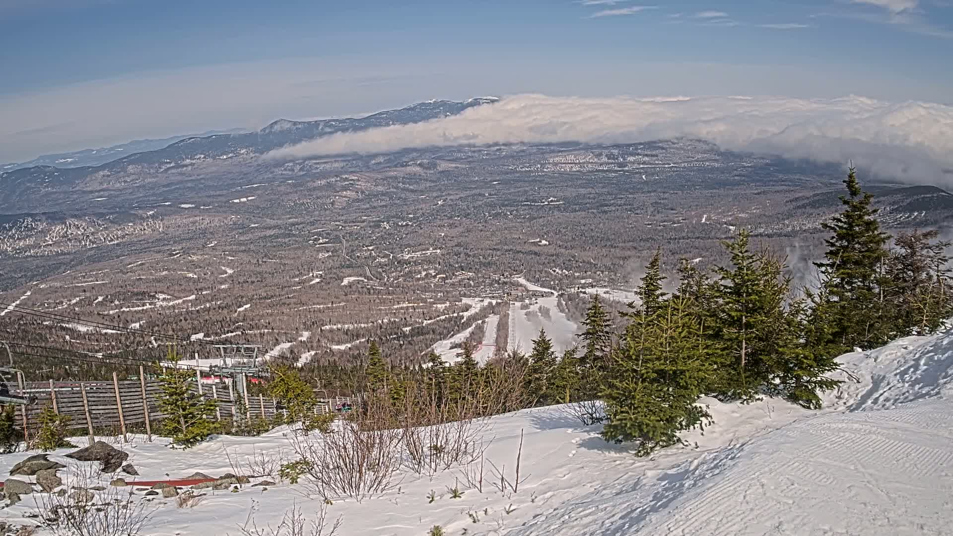 A snowy, mountainous landscape with evergreen trees in the foreground and a valley stretching to distant mountains partially obscured by clouds under a mostly clear blue sky.