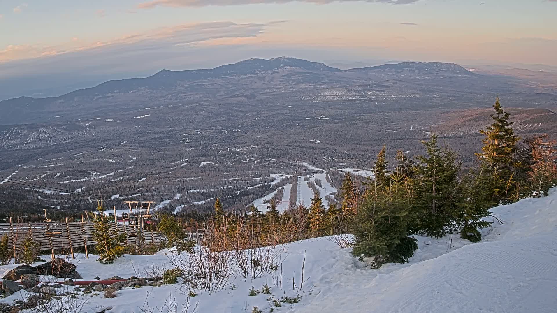 A snow-covered ski resort with a chairlift and expansive views of distant mountains under a partly cloudy, sunset sky.