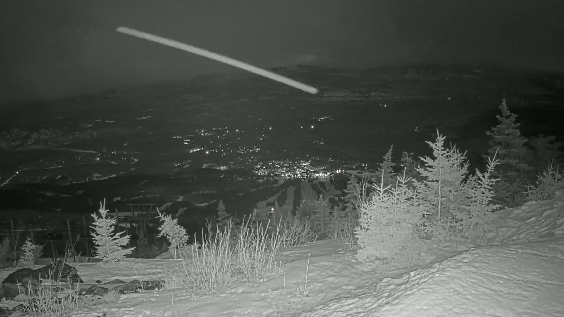 A nighttime, snowy landscape shows a meteor streaking across the sky above a valley with lights from a town below, viewed from a snow-covered hillside with evergreen trees.