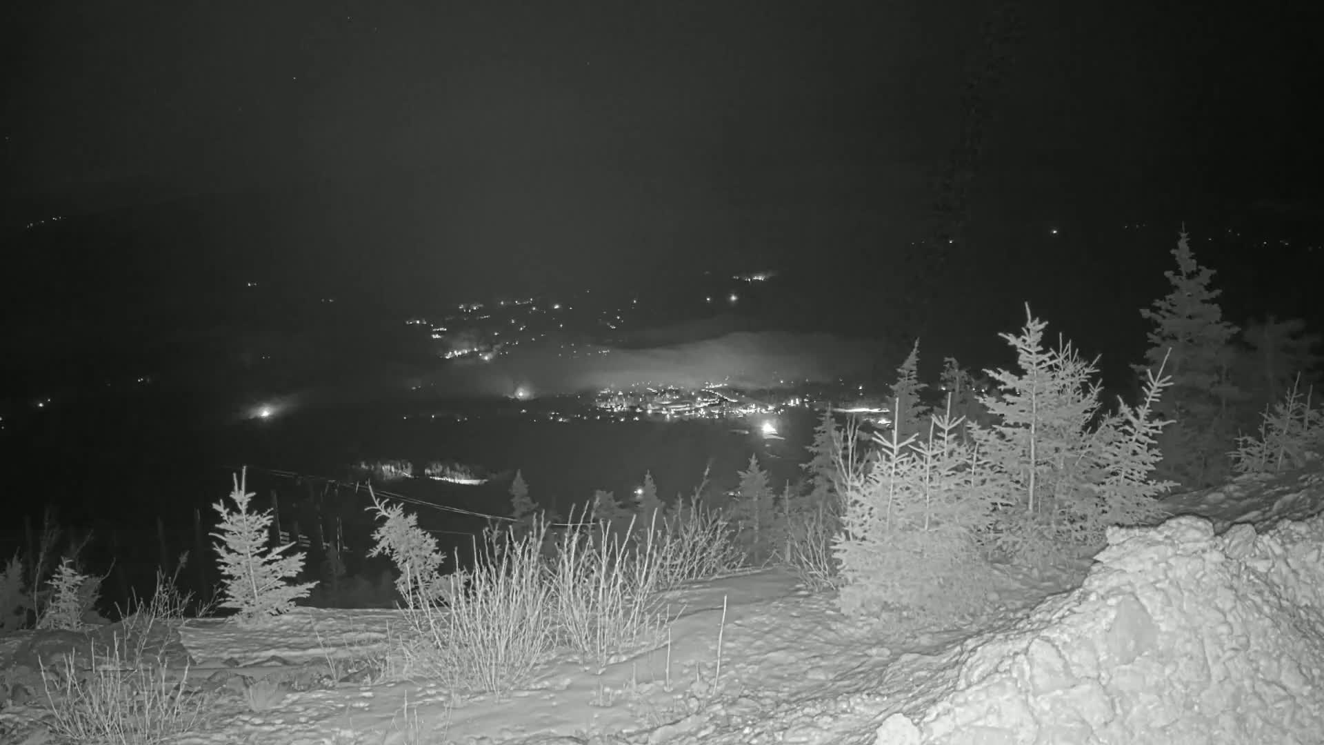 A nighttime view from a snow-covered hilltop shows a valley below with a town illuminated by lights and partially obscured by fog.