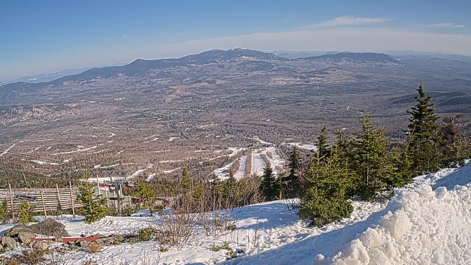 A snowy mountaintop view reveals a vast expanse of forested hills and valleys under a clear blue sky, with ski runs visible below.