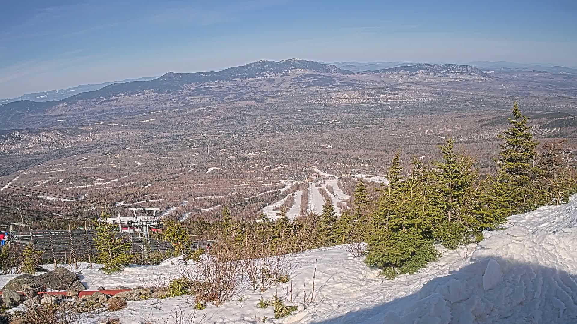 A snowy mountaintop view overlooks a vast forest and distant mountain range under a clear blue sky.