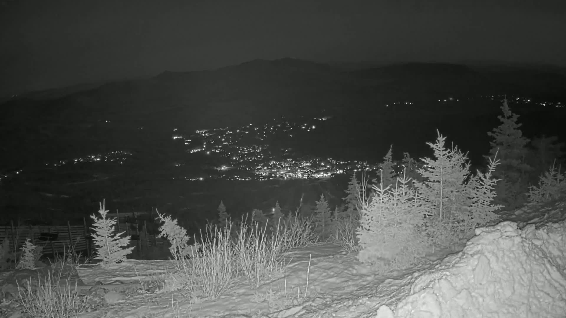A nighttime view from a snow-covered hilltop shows a valley with a town illuminated by lights below, under a dark sky.