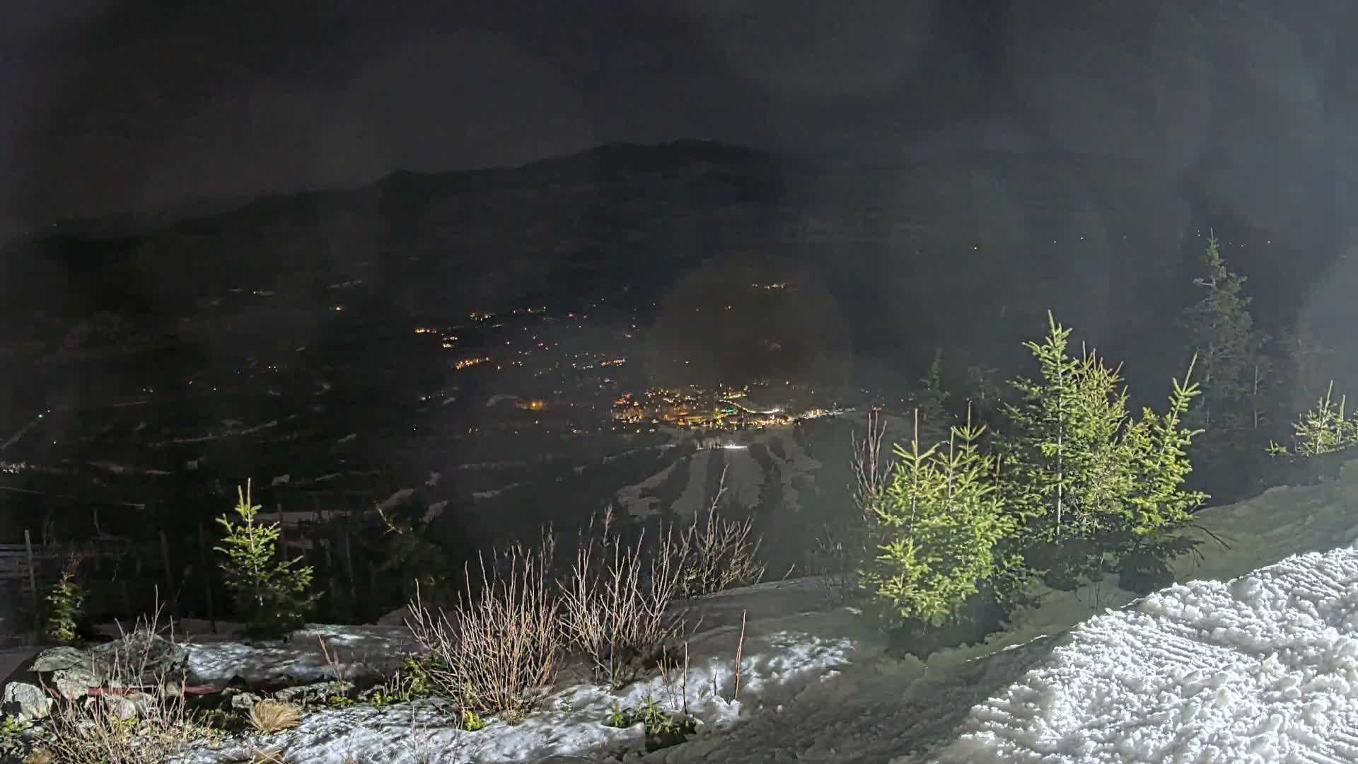 A nighttime view from a snowy mountaintop reveals a valley with a town illuminated below, under a hazy, dark sky.