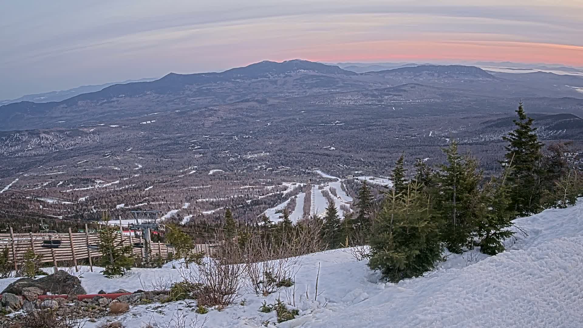 A snow-covered ski resort nestled in a vast, mountainous landscape under a pale pink sunset.