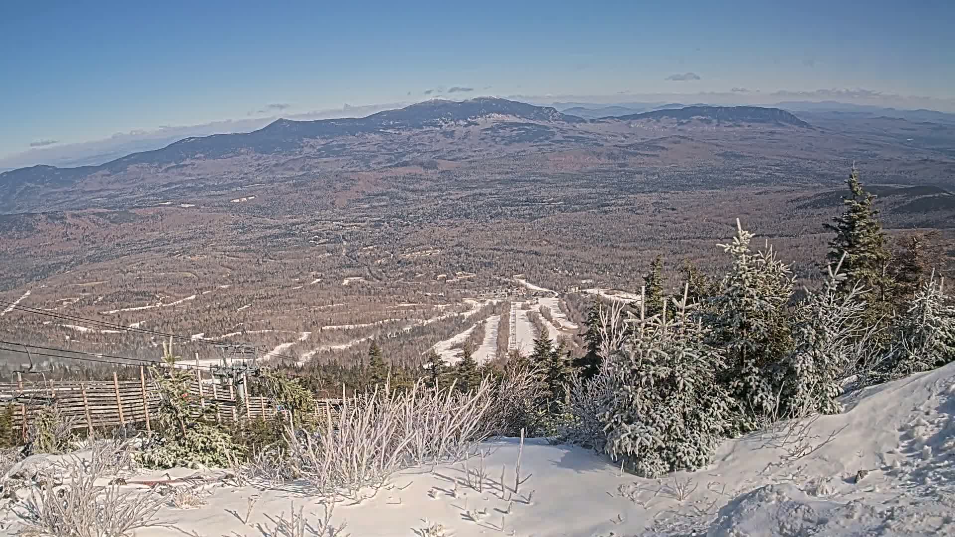 A snowy mountaintop view reveals a ski resort nestled in a vast, snow-dusted forest under a clear blue sky.